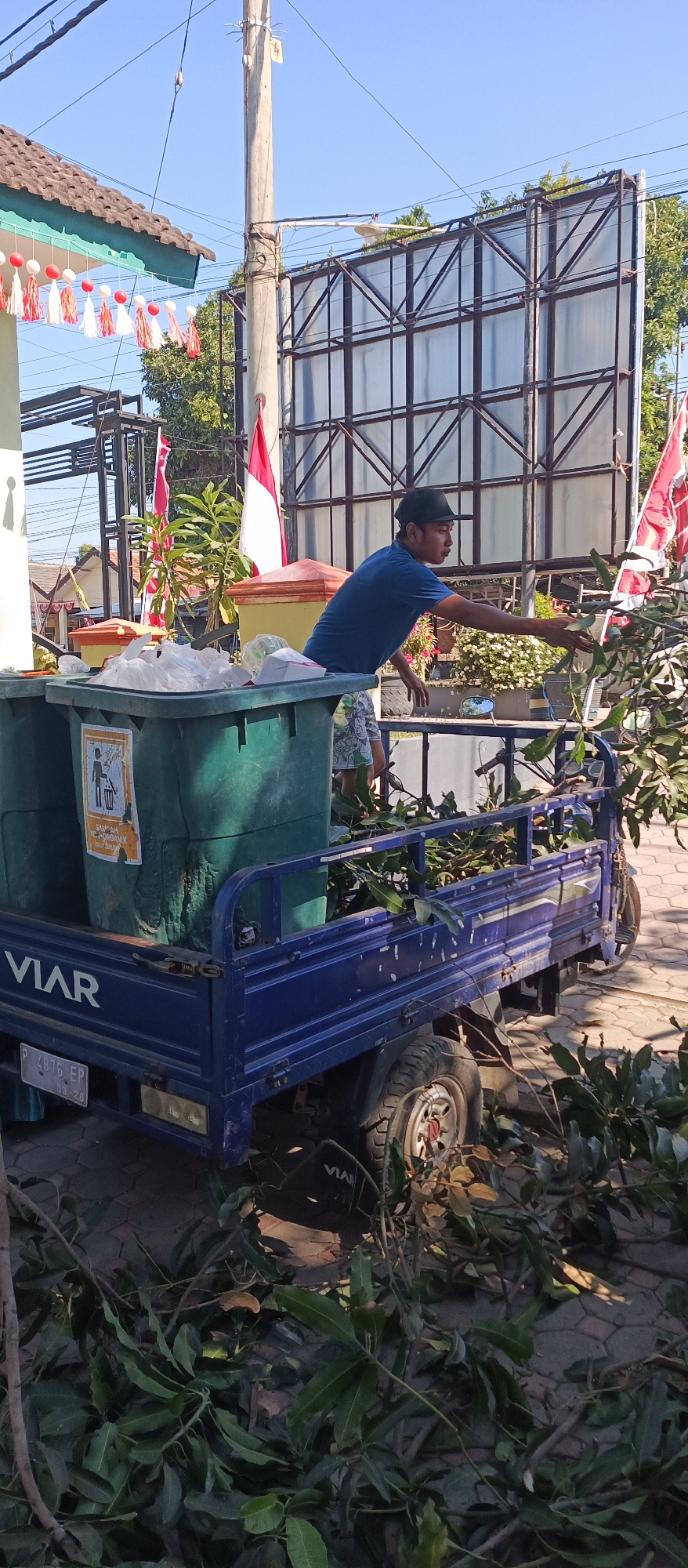 In Arjasa, Indonesia, a man wearing a blue shirt and a dark hat is seen leaning over a blue pickup truck. The truck is filled with green trash cans and leafy branches. The man appears to be reaching into the truck. The truck has "VIAR" written on its side. The scene is outdoors, with a blue sky and a large billboard structure visible in the background. There are also Indonesian flags and decorative tassels hanging from a building on the left side of the image. The ground is paved with interlocking bricks.