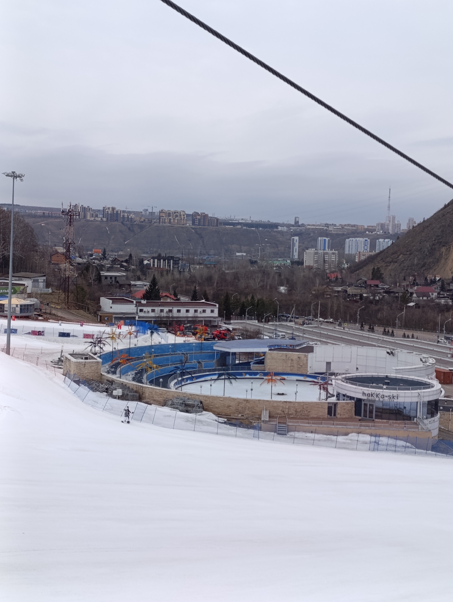 The image shows a winter sports scene in Krasnoyarsk, Russia, specifically the Bobrovy Log Funpark. A ski slope dominates the foreground, covered in snow. A lone skier is visible on the slope.
Behind the slope is a complex of structures, including an outdoor ice skating rink with decorative palm trees and a building labeled "hakKa-ski". These facilities are surrounded by a metal fence.
In the background, a cityscape stretches across the horizon, including residential buildings, industrial structures, and a tall communications tower. A hillside borders the city. The sky is overcast. A cable, likely part of a ski lift or aerial tramway, crosses the upper portion of the frame.