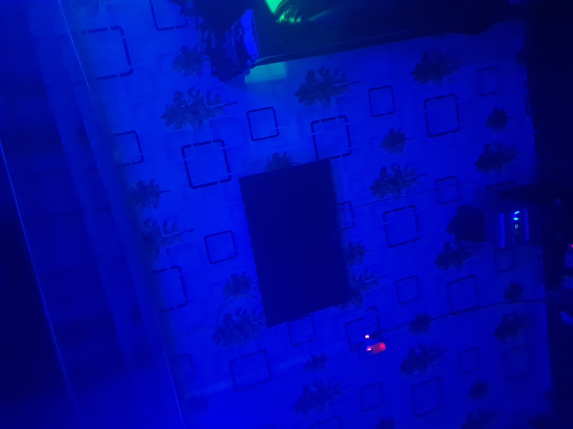 An interior scene, likely a room with blue ambient lighting, is captured from a low angle. The walls are decorated with a pattern of squares and stylized trees, creating a somewhat abstract or whimsical aesthetic. In the center of the image, a large, dark rectangular object is mounted on the wall. To the left, a small red light is visible, possibly indicating an indicator light or a small device. On the right side of the frame, a dark vertical object, possibly a pillar or a piece of furniture, is present, with some green light reflecting off its surface. The lighting conditions are dim and heavily tinted blue, which obscures fine details and adds a dramatic atmosphere to the scene. The floor area is largely in shadow and out of focus.