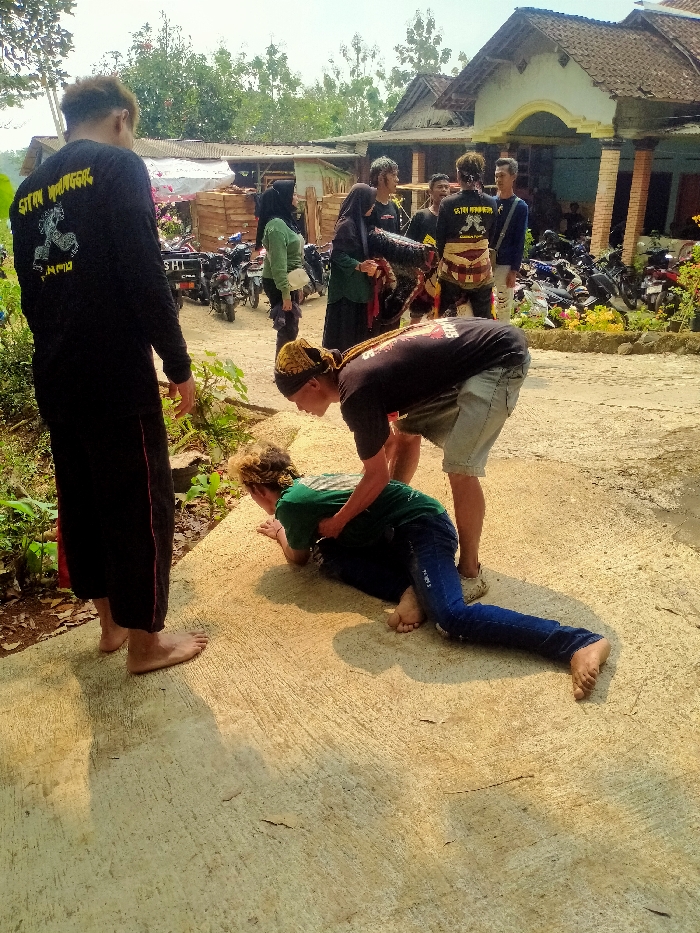 The image depicts an outdoor scene in Lempong, Indonesia, during daylight hours. In the foreground, two male individuals are engaged in a physical interaction on a light-colored concrete surface. One male, wearing a dark green short-sleeved shirt and blue jeans, is positioned on his hands and knees, with his head lowered. A second male, wearing a black t-shirt, light-colored shorts, and a brown and yellow patterned head covering, is bent over him, holding onto the back of his shirt. Both individuals are barefoot.
To the left of the central action, a third male stands facing away from the viewer. He is barefoot and wears a long-sleeved black shirt with the text "SATRIYA MANGKOK" and a horse-like logo printed in yellow on the back. His lower body is clad in loose black pants with a subtle red trim.
In the mid-ground and background, a group of approximately eight to ten additional individuals is observing the scene. This group includes both males and females; some males wear shirts similar to the one seen in the foreground, while some females wear hijabs. One male in the background wears a patterned traditional sash or belt. Behind these observers, a dirt path or unpaved area is visible, with multiple motorcycles parked. Further in the background, several simple residential structures are present, including one with a reddish-brown tiled roof and light-colored walls, and another with a corrugated metal roof and exposed wooden planks. Dense green vegetation and trees form the horizon. The sun is overhead, casting distinct shadows on the concrete surface.