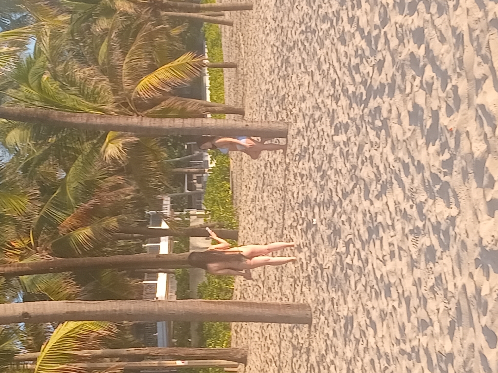 Two women are on a sandy beach, likely in Đà Nẵng, Vietnam, near palm trees. The woman on the left is facing away from the camera, wearing a brown bikini bottom. The woman on the right is also facing away, wearing a blue bikini. Both women appear to be standing. The background shows more palm trees and a building. The sand is lightly rippled, indicating it may be a beach after the tide has gone out.