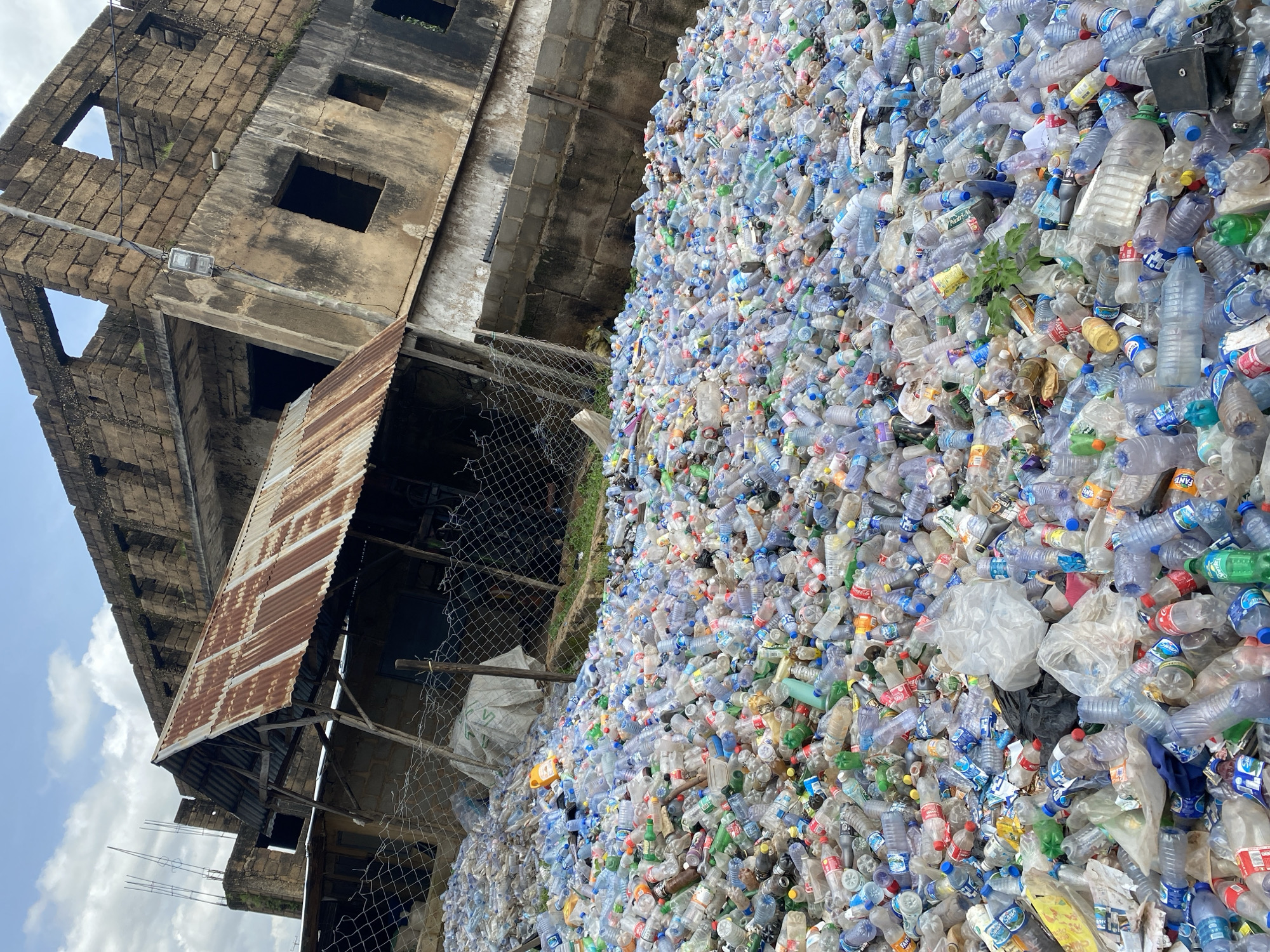 An outdoor scene in Gura Top, Nigeria, captures a vast accumulation of discarded plastic bottles filling the foreground and midground. The bottles, of various sizes, colors, and brands, are piled high, forming an expansive landscape of waste. In the background, a partially constructed or abandoned building looms, its weathered brickwork and gaping windows suggesting neglect. A makeshift structure with a rusted corrugated metal roof stands adjacent to the building, partially obscuring the view of its interior. A wire fence runs across the scene, separating the bottle-filled area from the building and the surrounding environment. The sky above is partly cloudy, with a mix of blue and white. The overall impression is one of environmental degradation and urban decay.