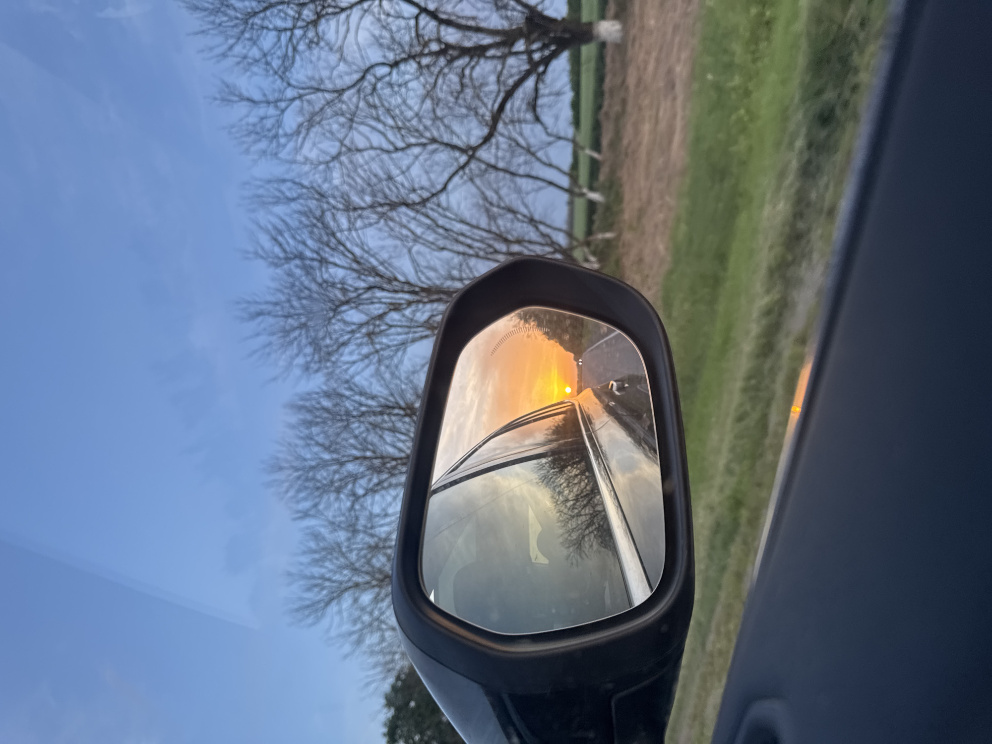 The image shows a car's side mirror reflecting a sunset over a road, viewed from inside the vehicle. The foreground includes the car's interior. The reflection shows a road receding into the distance, with the sun setting directly ahead. In the background outside the car, there are bare trees on the left and a field on the right. The photo appears to have been taken in the city of Novorozhdestvenskaya, Russia.