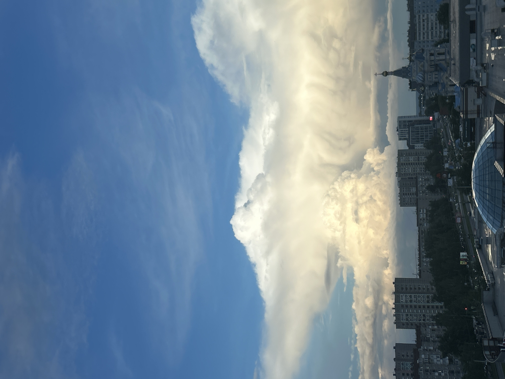 The horizontally oriented image captures a dynamic cityscape in Izhevsk, Russia, under a striking sky. The left portion of the sky is a vibrant, clear blue with faint, wispy cirrus clouds. This contrasts sharply with the right side, which is dominated by a massive, towering formation of cumulus clouds, brightly illuminated by the sun with shades of white and light golden yellow, suggesting either intense daylight or late afternoon sun.
Below the dramatic sky, an urban landscape unfolds. Prominently featured in the mid-right is a large church with multiple domes, one topped with a golden cross, characteristic of Russian Orthodox architecture. Surrounding this landmark are numerous multi-story buildings, including residential apartment blocks and other commercial or administrative structures. On the far right, a modern building with a distinct curved, glass facade is partially visible. Patches of trees and glimpses of streets are visible between the buildings, indicating a bustling urban environment. No specific human activity or identifiable people are visible, but the scene conveys a sense of a city experiencing rapidly changing weather conditions, with a clear blue sky juxtaposed against a grand, cloud-filled expanse. No discernible text is present.