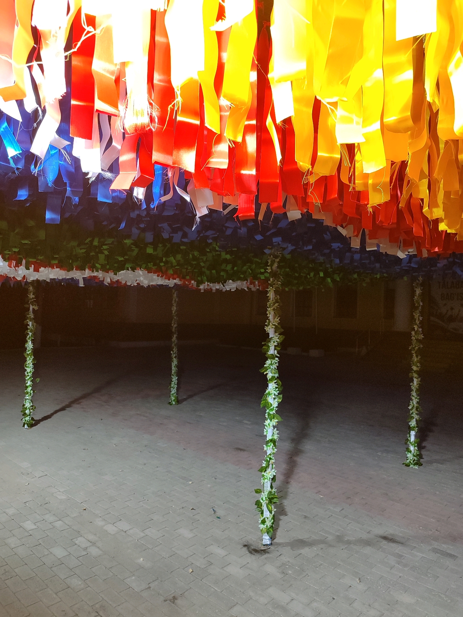 The image shows an outdoor scene at night, possibly in Nukus, Uzbekistan, featuring a colorful canopy of hanging ribbons in shades of yellow, orange, red, white, and blue. These ribbons are suspended from above, creating a decorative ceiling. Below the ribbon canopy, there is a band of green artificial foliage, with further decorative elements of white and red material. Supporting this structure are four vertical poles, two in the foreground and two further back, which are wrapped in artificial green vines and white flowers. The ground is paved with interlocking rectangular blocks. The overall impression is of an outdoor decoration or installation, possibly for an event.