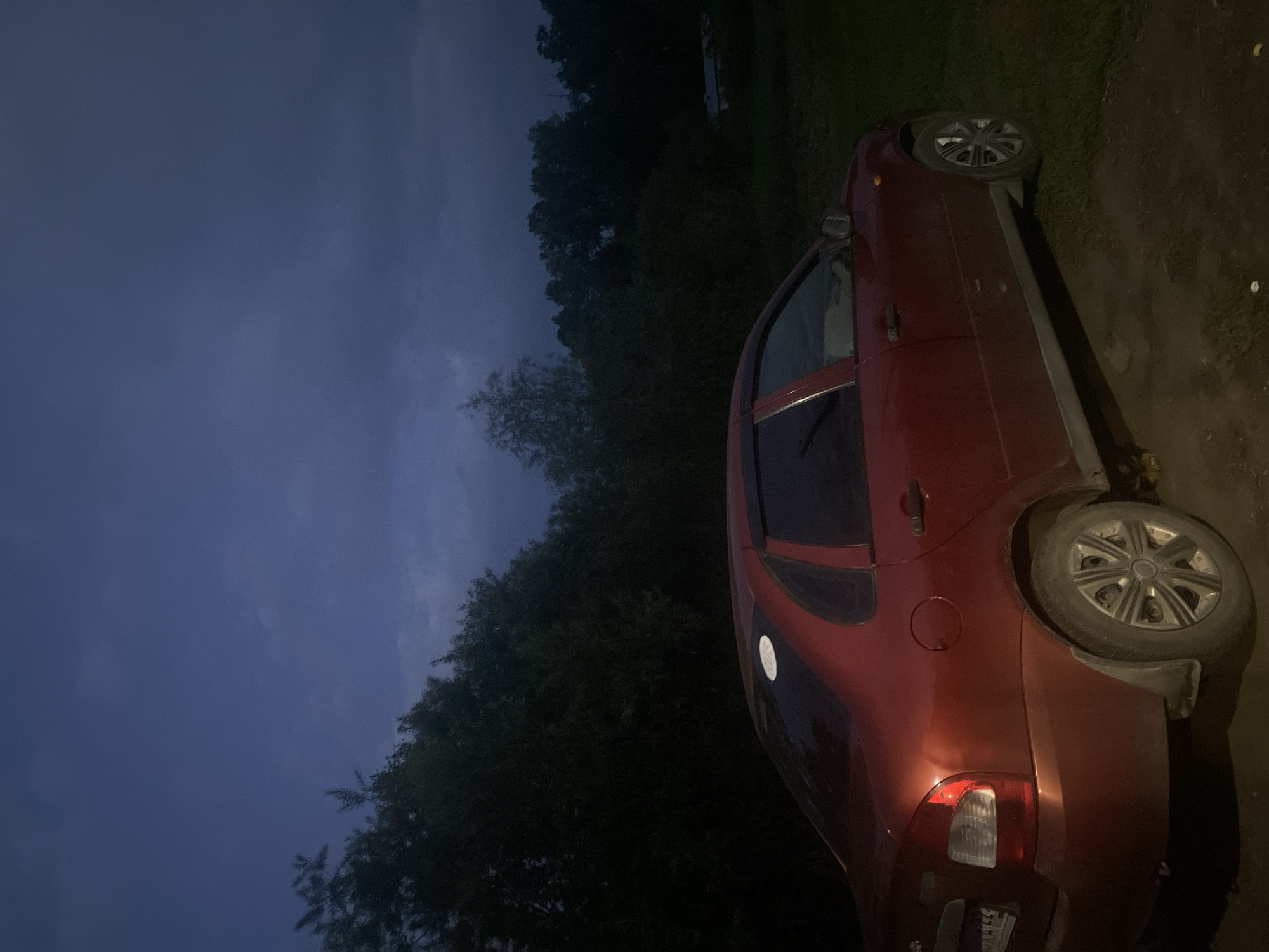 The image depicts a dark red sedan parked on an unpaved surface in a rural area. The car is positioned at an angle, with its rear left side closest to the viewer. The vehicle appears to be under a cloudy, twilight sky, with dense trees forming a dark backdrop behind it. To the right of the car, a patch of green grass is visible, and further in the background, a small building with a light-colored roof can be faintly discerned. The scene appears to be located in the village of Anton'yevka, Russia, given the details provided. There are no people visible in the image. The overall lighting suggests it is either early morning or late evening.