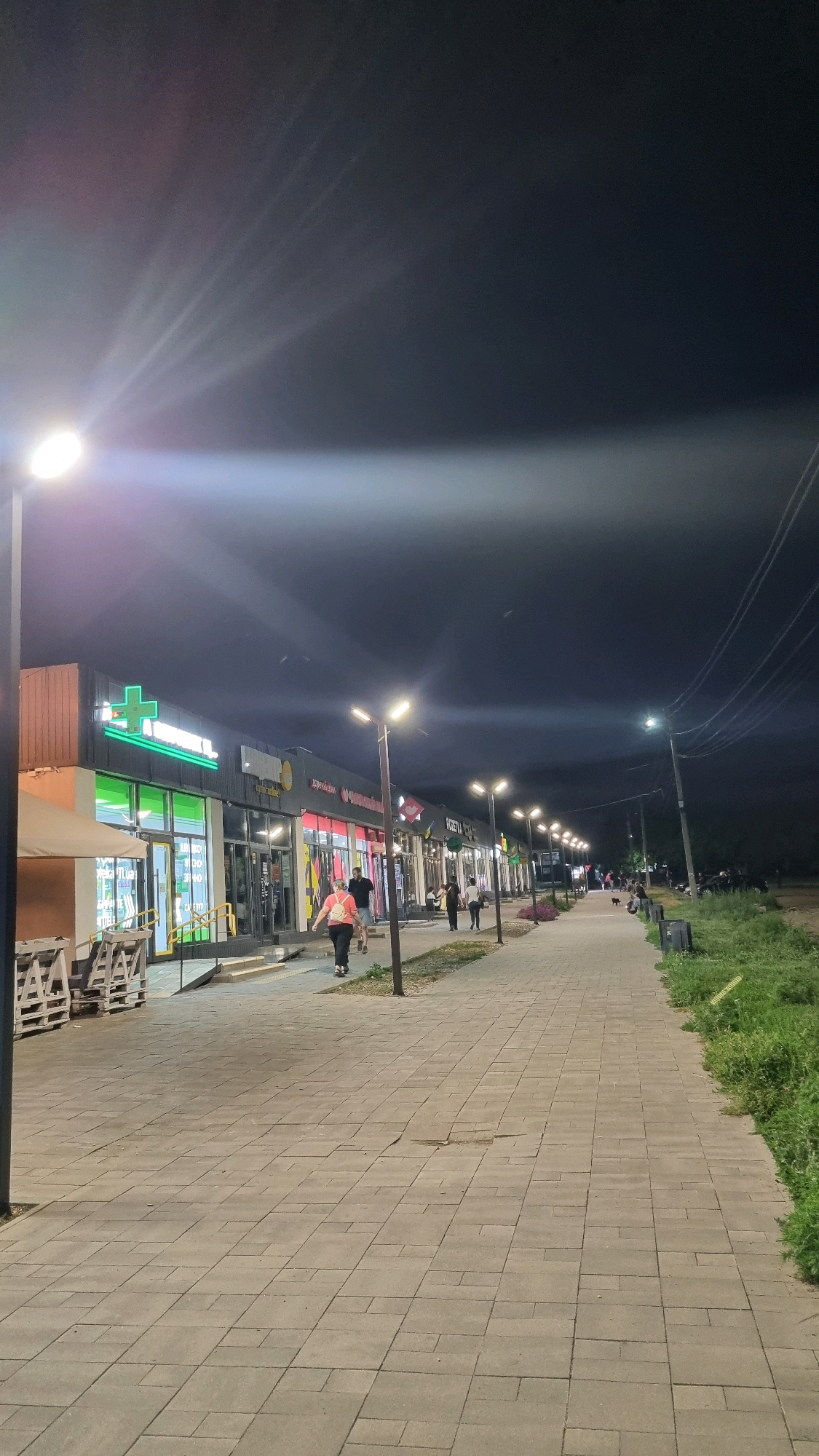 The image depicts a street scene at night in Smila, Ukraine. A paved sidewalk runs along a row of shops, illuminated by streetlights and the interior lights of the establishments. A woman in a coral-colored top and dark pants is walking on the sidewalk away from the camera. Other individuals are also visible walking along the sidewalk and near the shop entrances. The shops have large glass windows displaying merchandise, and some have illuminated signs, including one with a green cross. A large light fixture on a pole casts a strong beam of light across the upper portion of the image. To the right of the sidewalk, there is a grassy area with some bushes. The overall atmosphere is one of an active commercial street at night.