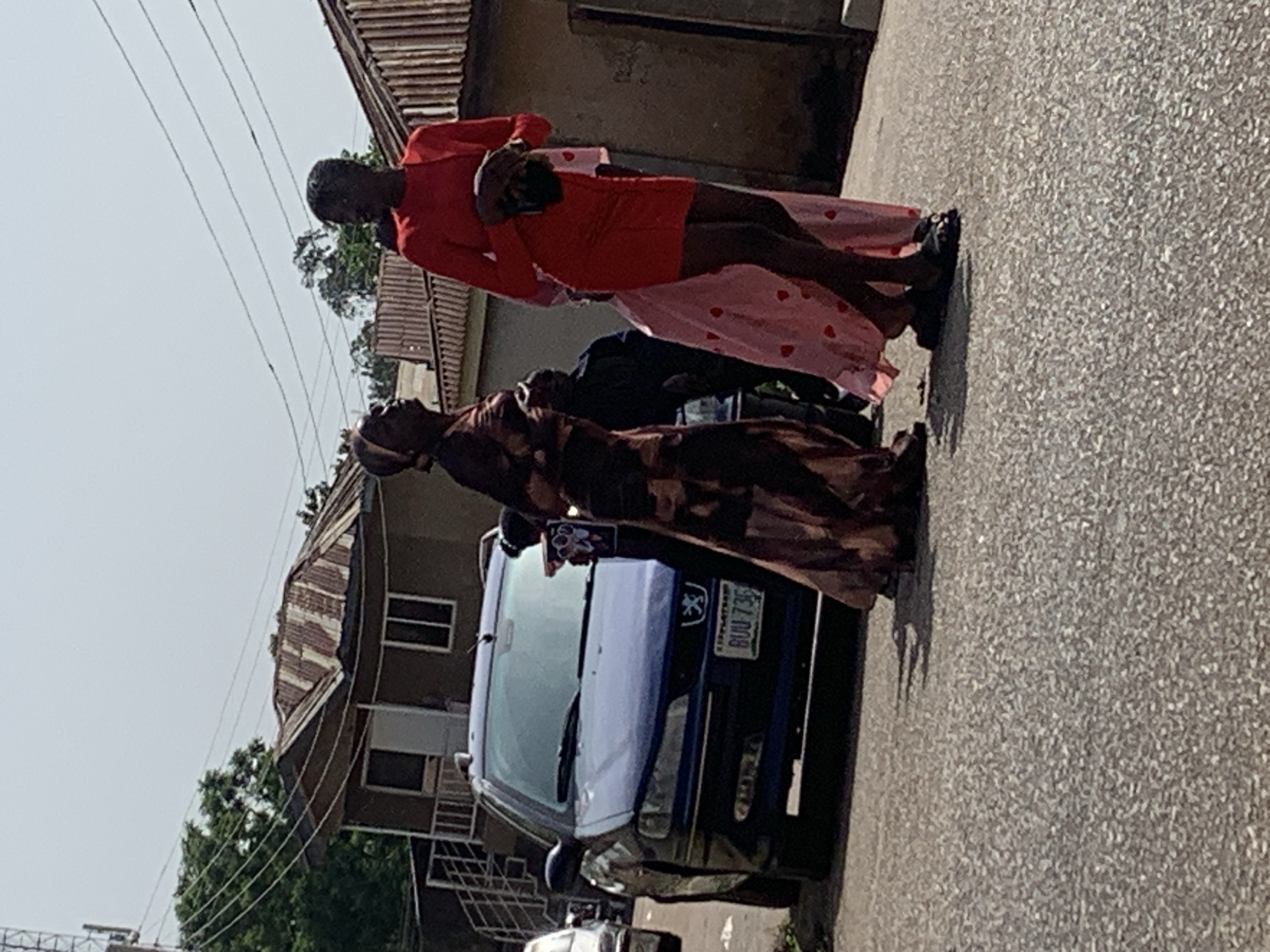The image depicts a street scene in Gura Top, Nigeria, with a blue Peugeot car parked on the right side of the asphalt road. Three individuals are visible. On the right, a person in a red dress stands with a dark clutch bag. To their left, a person in a brown and patterned dress walks, wearing a headscarf. Partially visible between them is a person wearing a pink dress with heart patterns, and another individual in a dark top. The background shows buildings with corrugated iron roofs, trees, and utility poles with wires. The sky is overcast. The license plate of the car is visible, reading "BUU 73G".