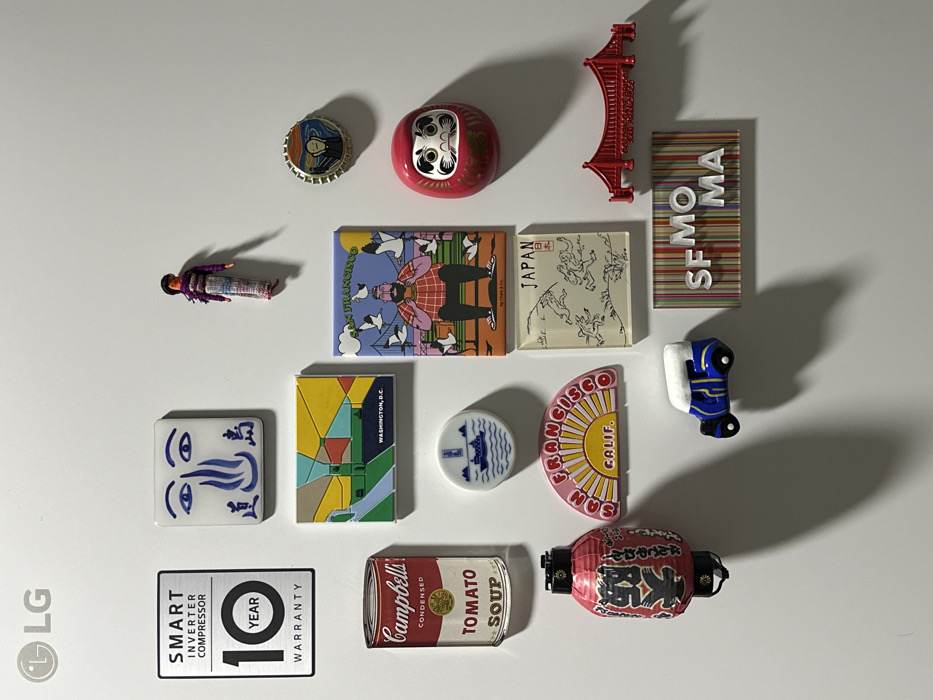 The image displays a collection of sixteen distinct magnets and stickers affixed to a vertical, smooth white surface, consistent with a refrigerator door. The surface is identified by an LG logo sticker in the top left corner and a "SMART INVERTER COMPRESSOR 10 YEAR WARRANTY" sticker directly below it. The scene is located in Mexico City, Mexico.
From top-left, the arrangement includes:
1. An LG logo sticker.
2. A silver rectangular sticker advertising a "SMART INVERTER COMPRESSOR 10 YEAR WARRANTY".
3. A white square magnet featuring a blue abstract face or calligraphy design.
4. A small, multicolored woven doll magnet.
Below these are:
5. A rectangular magnet with a colorful, geometric depiction of a city, labeled "WASHINGTON, D.C.".
6. A can-shaped magnet resembling a "Campbell's Condensed TOMATO SOUP" can.
7. A rectangular magnet illustrating a bearded man surrounded by seagulls, labeled "SAN FRANCISCO" and "by Chato & Co.".
To the right, positioned higher than the Washington, D.C. magnet:
8. A red bottle cap-shaped magnet featuring Edvard Munch's "The Scream" artwork.
9. A red Daruma doll-shaped magnet, positioned below the bottle cap.
Mid-lower section of the surface features:
10. A small, circular white magnet with a blue ship design.
11. A red lantern-shaped magnet with black Japanese characters.
12. A semi-circular magnet with a yellow sunburst design, labeled "SAN FRANCISCO CALIF.".
13. A clear rectangular magnet with black line drawings of figures and the word "JAPAN".
14. A red plastic Golden Gate Bridge-shaped magnet.
At the bottom of the collection are:
15. A blue and white cartoon-style car magnet.
16. A rectangular magnet with vertical multicolored stripes and the white letters "SFMOMA".
All items are static and adhere to the white surface, casting shadows consistently from an upper-left light source, indicating their three-dimensional form. No individuals are present in the immediate environment.