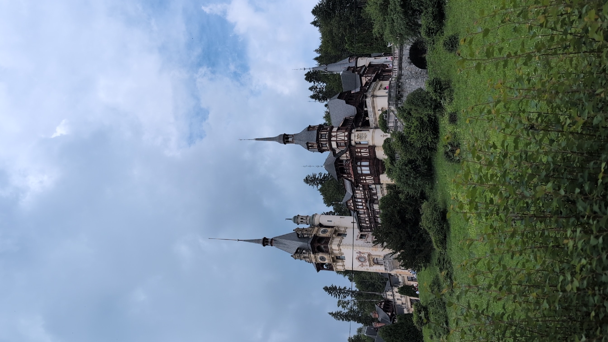 This media file depicts Peles Castle in Sinaia, Romania, during daytime with overcast skies. The castle, a grand architectural marvel, is surrounded by lush green foliage and mature trees. Its distinctive turrets and intricate half-timbered facade are prominent features. The image shows a side view of a section of the castle, highlighting its historical and ornate design. The castle appears to be situated on a slope, with the grounds descending into greenery.
The scene suggests a leisurely visit to a historical landmark, with the overcast weather contributing to a serene and slightly muted atmosphere. The presence of trees and the expansive grounds indicate a natural and picturesque setting, likely a park or garden area surrounding the castle. No people are clearly visible, but the general ambiance implies a tourist destination. The overall impression is one of historical grandeur set within a tranquil, natural environment. The image is oriented vertically, requiring rotation for a natural perspective of the castle.