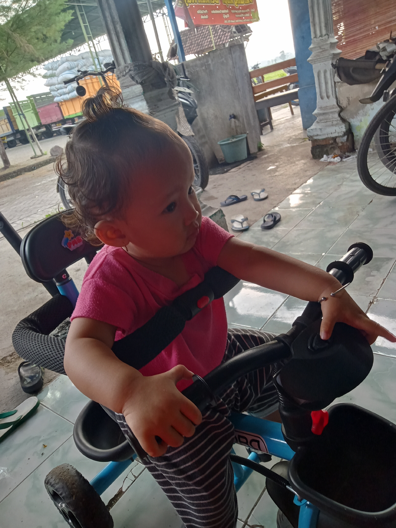 A young child with curly hair pulled up in a small bun is seated on a blue tricycle with a black seat and handlebars. The child is wearing a pink shirt and striped pants. They are holding onto the handlebars with both hands.
The tricycle is on a tiled surface with flip-flops scattered nearby. In the background, there is a building supported by pillars, motorcycles, and a truck loaded with large sacks under a metal structure. A bench is visible in the background as well. The image was taken outside during daylight hours in the city of Bungur, Indonesia.
