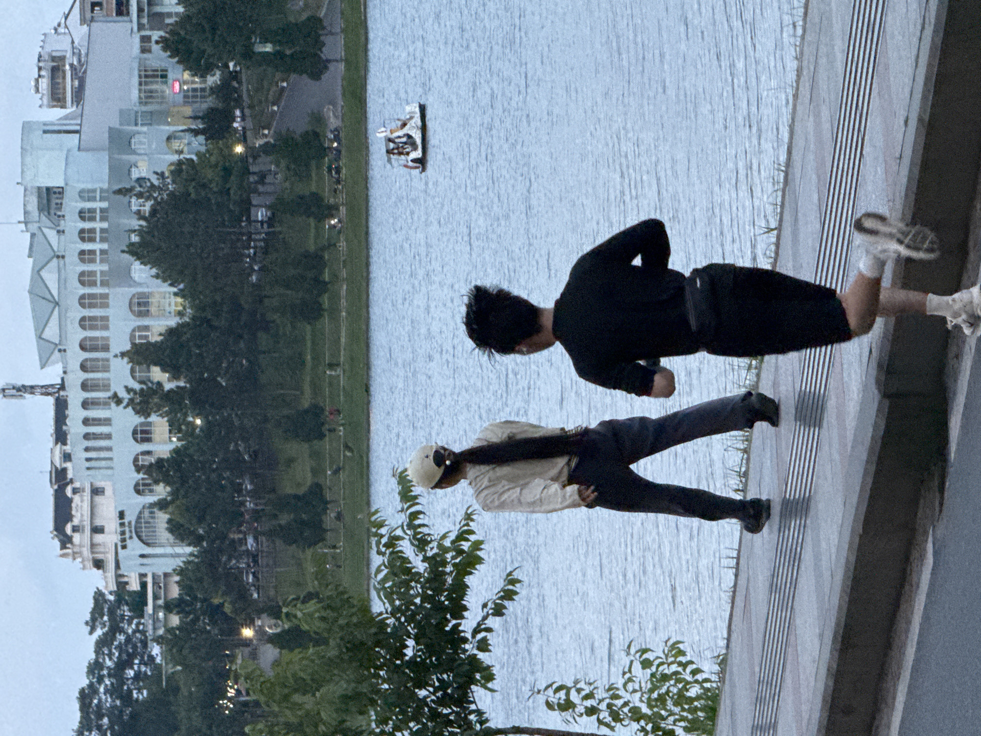 The image depicts a lakeside scene in Xuan Huong Lake, Da Lat, Vietnam. In the foreground, two individuals are engaged in physical activity. A person in the foreground is running away from the camera, wearing a black long-sleeved shirt, black shorts, white socks, and white sneakers. They have a fanny pack around their waist. To the left, another individual is walking away from the camera, wearing a light-colored long-sleeved shirt, dark pants, and a light-colored cap. Their long dark hair is visible down their back.
The background features a body of water, presumably Xuan Huong Lake, with a small pedal boat carrying at least three people in the distance. Beyond the lake, a tree-lined area leads up to a collection of buildings. One prominent building is light blue with arched windows and appears to be a large structure, possibly a hotel or a public building. Other buildings with a mix of architectural styles, including white facades and dark roofs, are visible further in the background, interspersed with trees. The overall lighting suggests it is either early morning or late evening. The ground in the immediate foreground is a paved walkway with a textured surface.