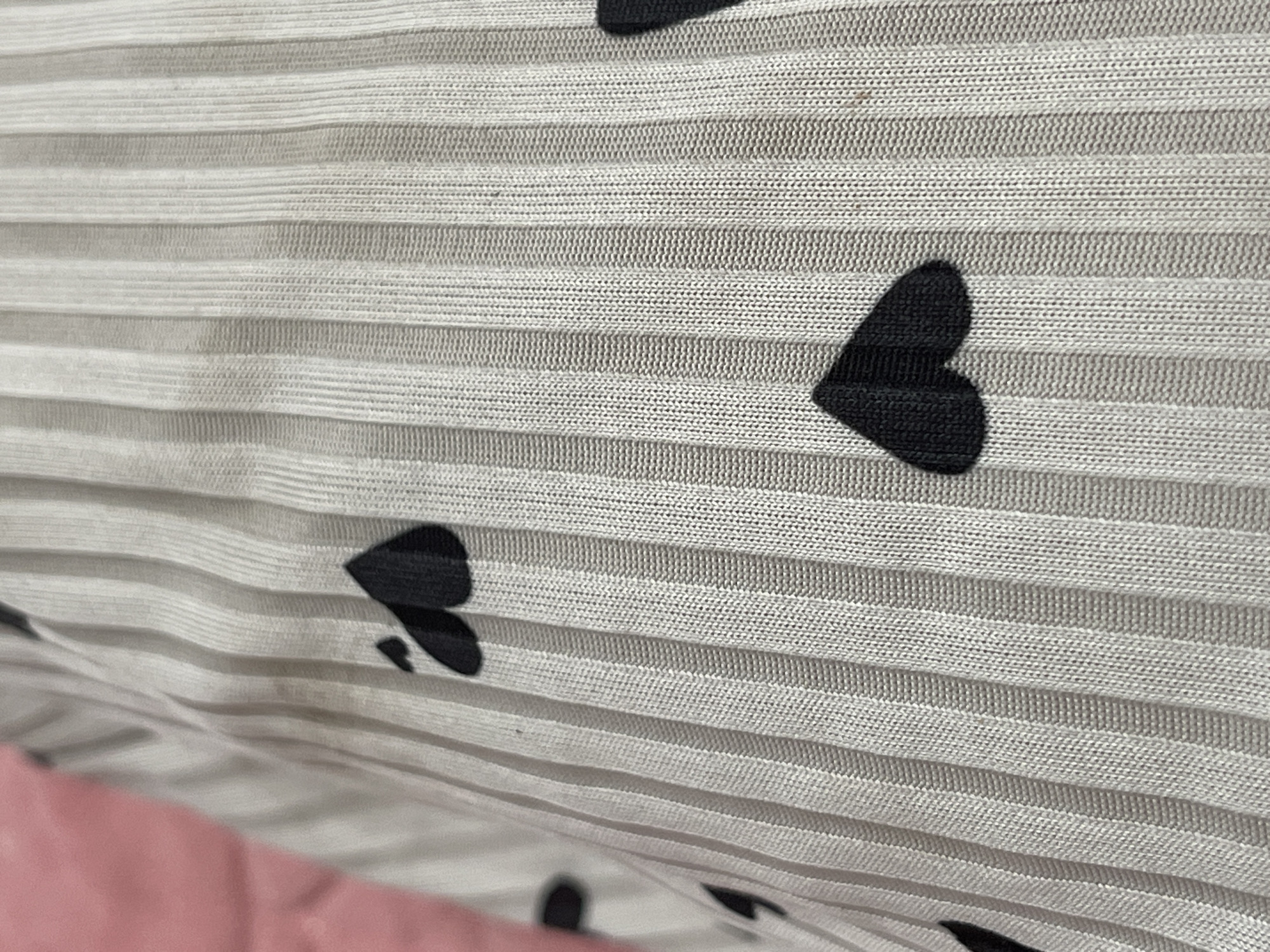 The image shows a close-up view of a light-colored fabric with a ribbed texture, featuring a pattern of black heart shapes. The fabric appears to be a garment, possibly a t-shirt or sweater, given the ribbed knit and the playful heart motif. The background is out of focus, with a pinkish hue visible on the left side, suggesting it might be another piece of clothing or a soft surface. The lighting is soft, casting subtle shadows that highlight the texture of the fabric. There are no identifiable landmarks or elements that would place this image specifically in Bishkek City, Kyrgyzstan. The focus is entirely on the textile and its pattern.