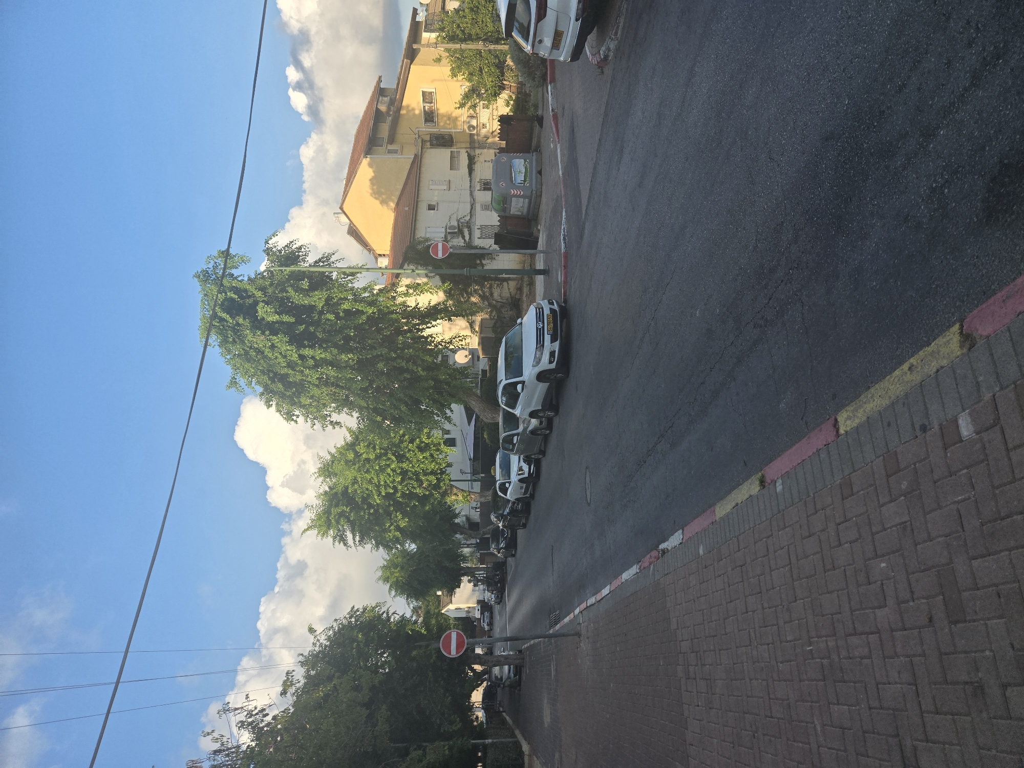 The image captures a street scene in Herzliya, Israel, likely during the daytime with some clouds present in a blue sky. The perspective is from a sidewalk, looking down a paved road lined with parked cars. A sidewalk made of reddish-brown brick pavers runs along the left side of the road, bordered by a curb with red and white stripes, and further by a yellow stripe.
On the right side of the road, several cars are parked. The most prominent are a white Volkswagen SUV and a white Kia SUV. Behind them, other vehicles are visible. The road itself is asphalt and shows signs of wear.
Lining the street are numerous trees with lush green foliage, casting some shadows. In the background, there are buildings with light yellow and orange facades, featuring red tiled roofs. One building appears to be multi-story. A few air conditioning units are visible on one of the buildings.
A red and white circular "no entry" sign on a pole is prominently positioned on the left side of the road, near the intersection. A large green dumpster with some graphics is situated near the buildings on the right side of the road. Overhead, power lines stretch across the sky. No people are visible in the image. The overall impression is of a residential or commercial street in a Mediterranean climate.