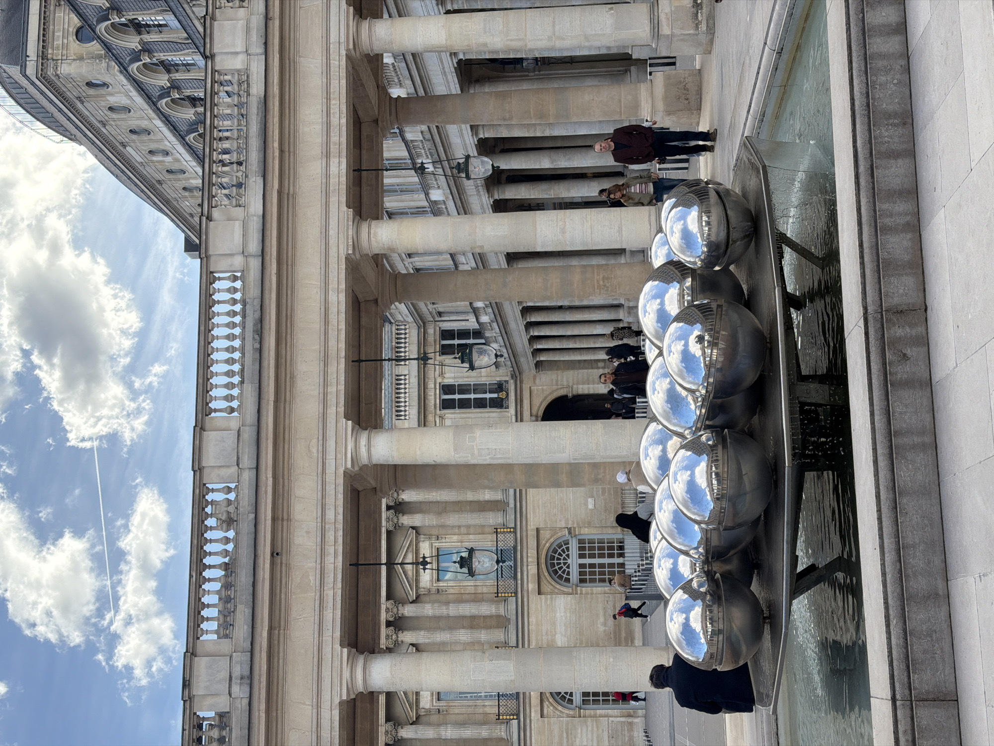 The scene depicts the Cour d'Honneur of the Palais-Royal in Paris, France, under a partially cloudy blue sky with a distinct aircraft contrail in the upper left. In the immediate foreground, a rectangular stone fountain basin contains a modern art installation: a collection of approximately 15 large, highly reflective spherical metal orbs arranged on a dark, multi-tiered platform. Water flows from the platform into the basin, with the orbs reflecting the sky and surrounding classical architecture.
Behind the fountain, a grand colonnaded walkway stretches horizontally, composed of numerous tall, light-colored stone columns with classical capitals supporting an entablature and a balustrade above. Several ornate dark metal lanterns are suspended from the colonnade's ceiling. Beyond this, the classical architecture of the Palais-Royal complex continues, presenting multiple stories of light-colored stone facades featuring arched and rectangular windows. A curved, dark-tiled roof section with circular and arched dormer windows is visible in the upper right background.
Several individuals are present. In the foreground left, a person wearing a dark coat is seated on a stone ledge adjacent to the fountain, viewed from behind. In the midground, a man in a maroon jacket and dark pants stands on the right, facing slightly towards the left. A woman in a green jacket and striped shirt stands to his left. Other individuals, including a person with a dark head covering and another wearing a light-colored beanie, are visible near the fountain. Additional figures are sparsely distributed throughout the background, either walking or standing within the courtyard and under the colonnades. No specific actions or interactions beyond general movement and observation are discernible among the individuals.