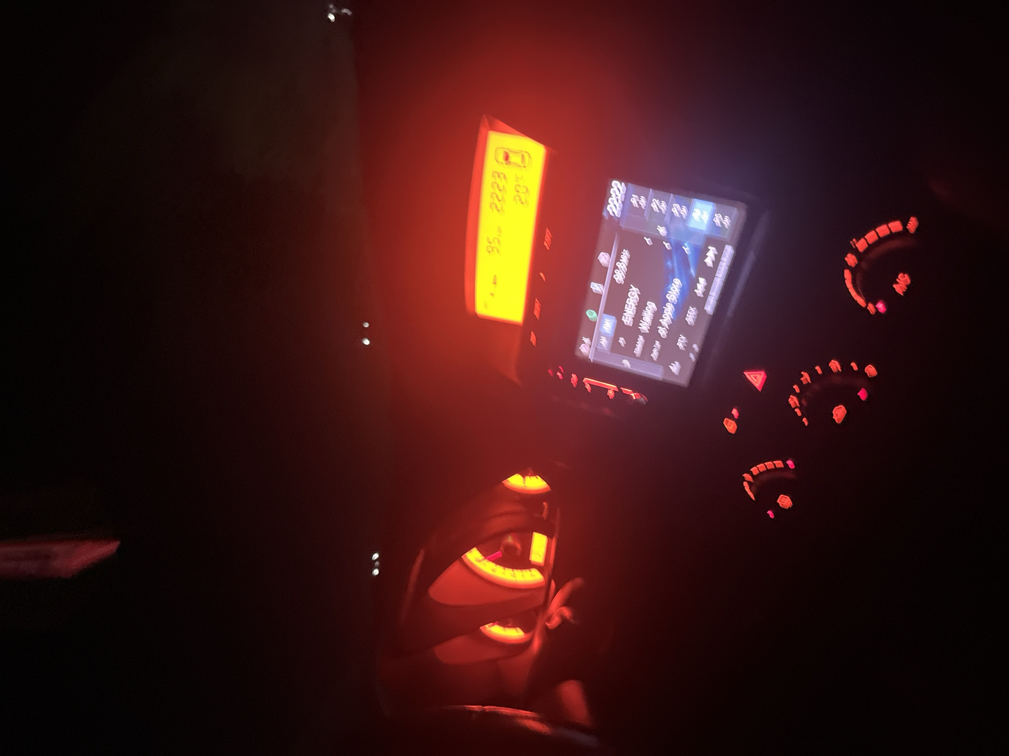 The image displays the interior of a vehicle at night, characterized by the prominent red and orange illumination of its dashboard and central console. The scene is set inside a car, with the location context being Blagoevgrad, Bulgaria.
On the left, a rectangular digital display provides vehicle information. It shows the numbers "95" and "2223", with a car icon and "20" visible beneath them, likely indicating a percentage such as battery level or fuel efficiency.
To the right, the vehicle's infotainment system is active, featuring a screen that prominently displays "22:22", indicating the time is 10:22 PM. Below this, "88.8MHz" suggests a radio station is currently selected. Other visible text on the screen includes "ENERGY", "Waiting", and "Apple Store". Accompanying buttons are labeled "AM", "FM", "SIRIUSXM", "TUNE", "SEEK", and "Pty".
Further to the right, a set of circular controls, presumably for the climate system, are illuminated in a vibrant orange-red. The letters "ANS" are visible on one of these controls. The lower part of the image reveals sections of the steering wheel or central console and what appears to be a gear shift lever, all contributing to the ambient red glow within the cabin.
No people are visible in the image, which captures the functional and illuminated interior of a vehicle operating during nighttime.