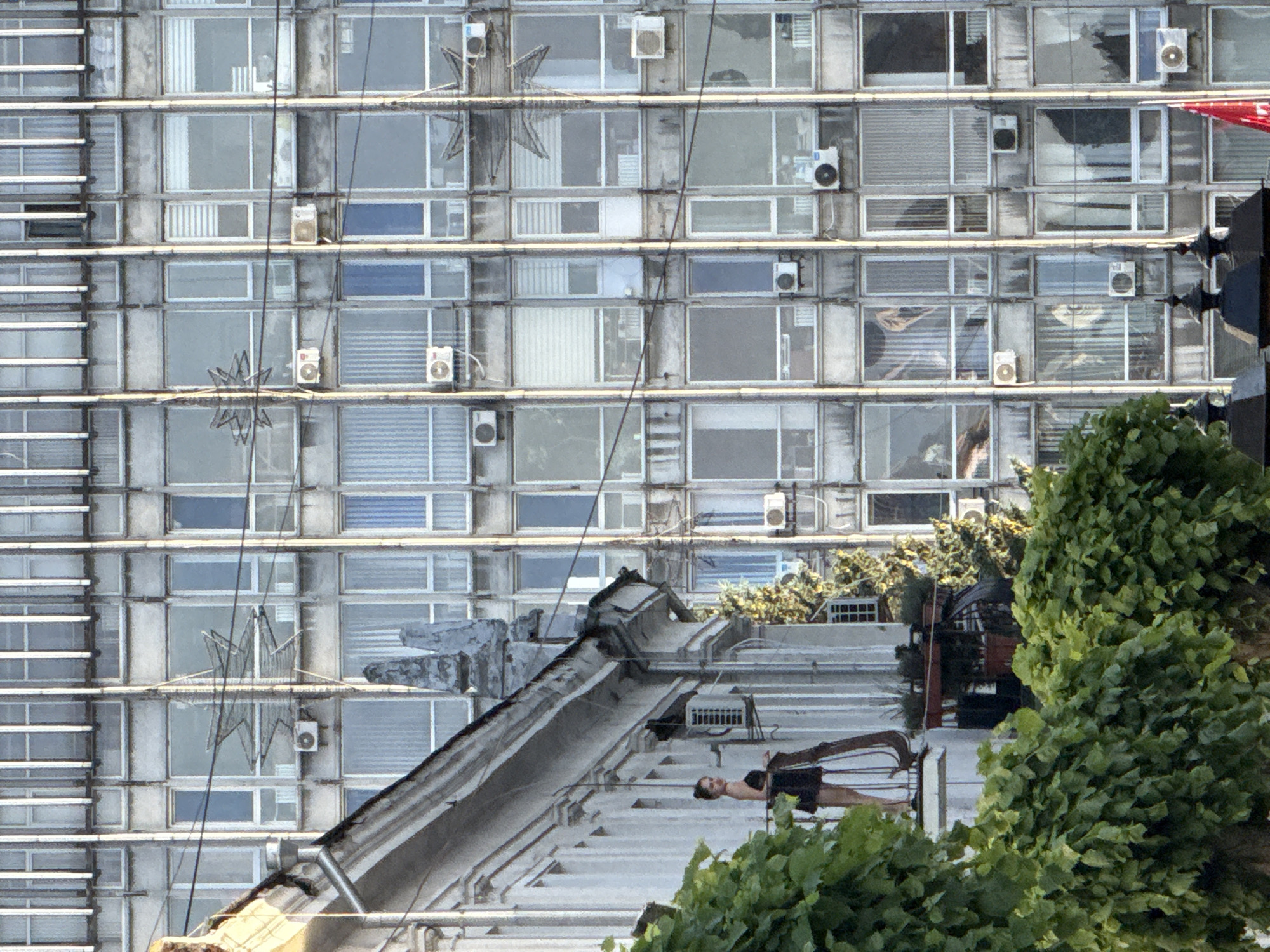 The image depicts a woman standing on a balcony of a building in Vinnytsia, Ukraine. She is wearing a black dress and has her hair pulled back. The balcony is partially obscured by green foliage from trees. Behind the woman and her building, a large, gray, modern building with numerous windows is visible. Some of these windows have air conditioning units installed. The architecture of both buildings suggests an urban environment. The overall scene captures a moment of stillness on a balcony against the backdrop of a prominent city structure.