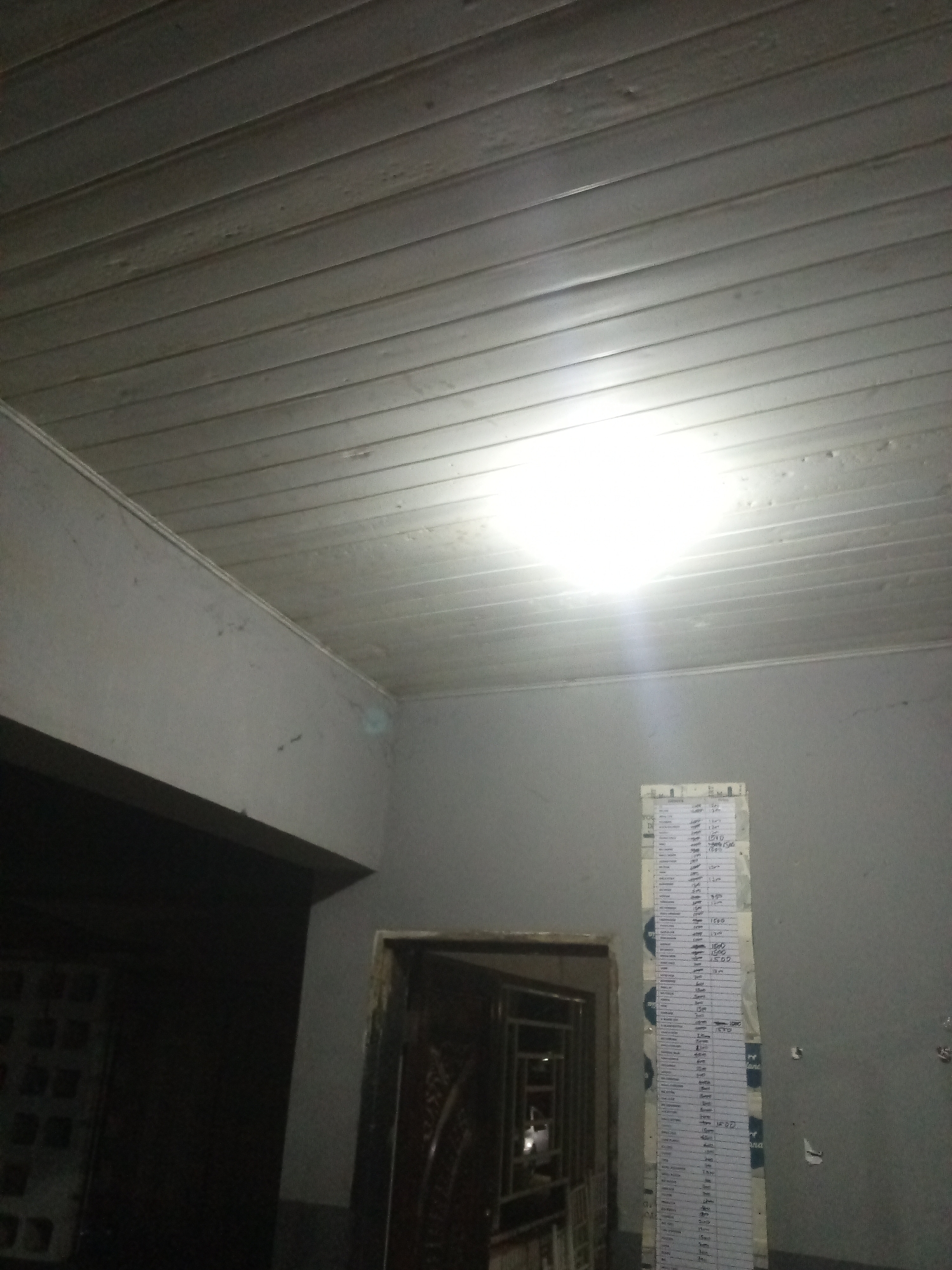 A tall, vertical sheet of paper with rows of text and numbers is taped to a gray wall in an indoor location. The paper appears to be a list or a table, with handwriting visible in blue ink. The wall is plain and shows some minor imperfections. Above the wall is a white, slatted ceiling, illuminated by a bright overhead light. To the left of the wall with the paper is a dark doorway with what appears to be a decorative door. To the right of the paper, another doorway or opening is visible. The overall impression is of a dimly lit room, possibly a hallway or a storage area, with a functional, unadorned aesthetic.