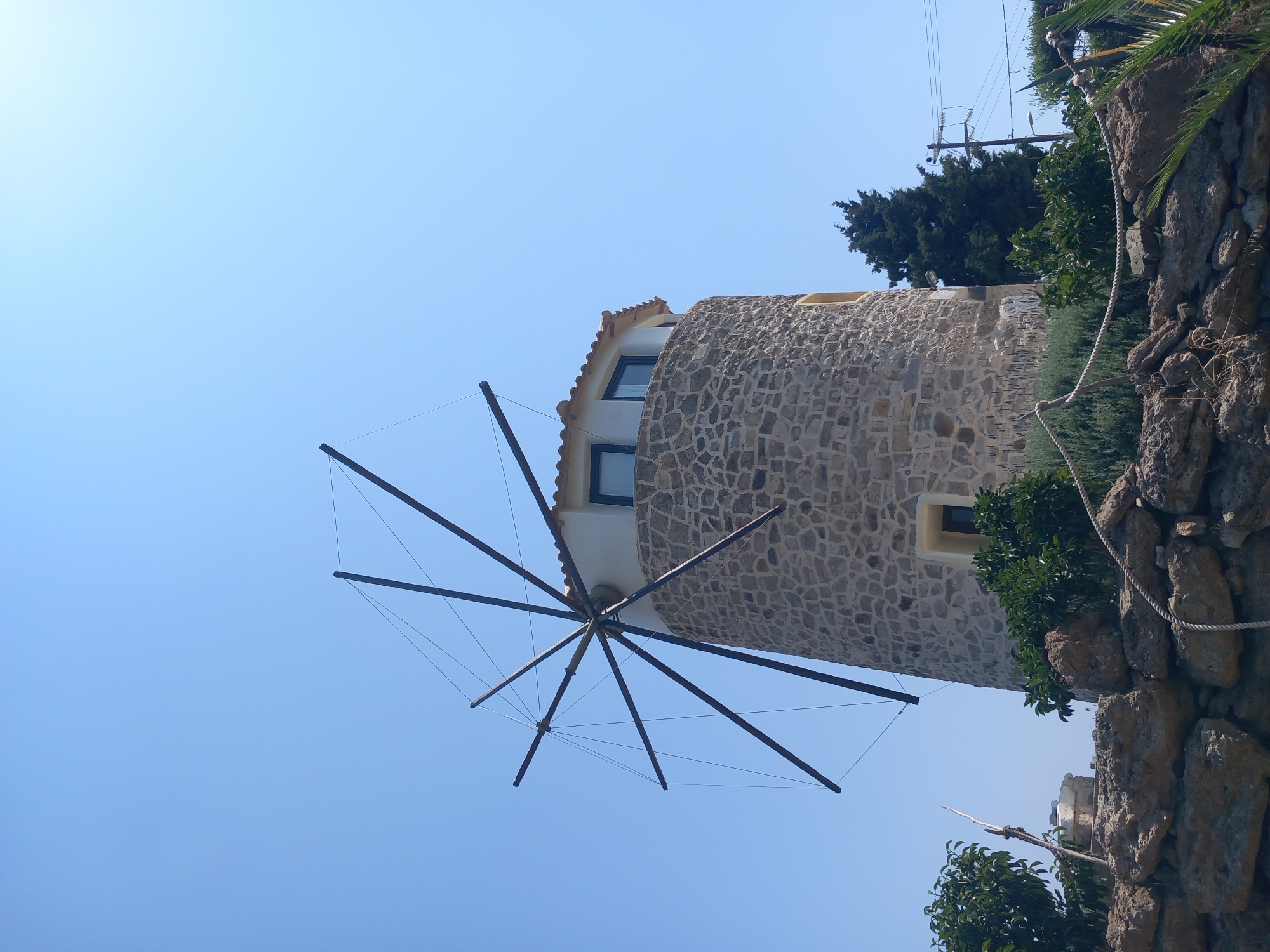 The image shows a stone windmill in Platani, Greece. The windmill has a conical shape and is constructed from rough-hewn stones. It has several windows and a tiled roof. The windmill's sails are visible against a clear blue sky. In the foreground, there is a low stone wall, and behind it, there are various plants and trees, including a tall evergreen tree. A rope is strung along the top of the stone wall. The overall impression is one of a rustic, historical structure set within a natural landscape.