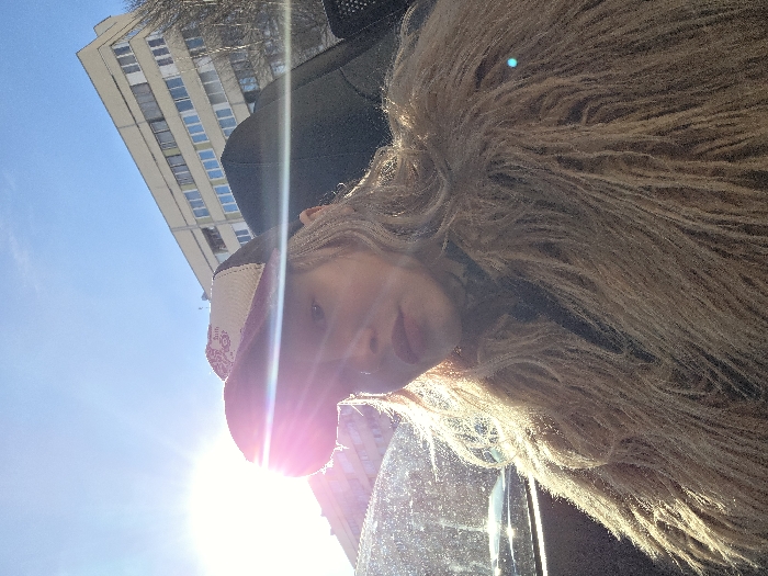 A female individual is depicted in a close-up, upward-angled self-portrait, seated within what appears to be a vehicle. She has long, wavy blonde hair, partially covered by a two-toned baseball cap with a dark brim and a lighter, patterned crown. Her lips are prominent and appear to be enhanced with makeup. She wears a thick, shaggy, light-brown faux fur coat.
The scene is brightly illuminated by direct sunlight originating from the upper left, causing a significant lens flare across the subject's face and the upper portion of the image. The sky is clear and blue. In the background, a multi-story, light-colored building with multiple rectangular windows is visible. Its architecture is typical of mid-to-late 20th-century urban development. Bare tree branches are visible to the right of this building, suggesting a cold season. The general urban environment is consistent with areas in Saint Petersburg, Russia, but specific landmarks or unique architectural features required for precise location identification within the city are not visible. No interactions with other individuals or objects are occurring.
