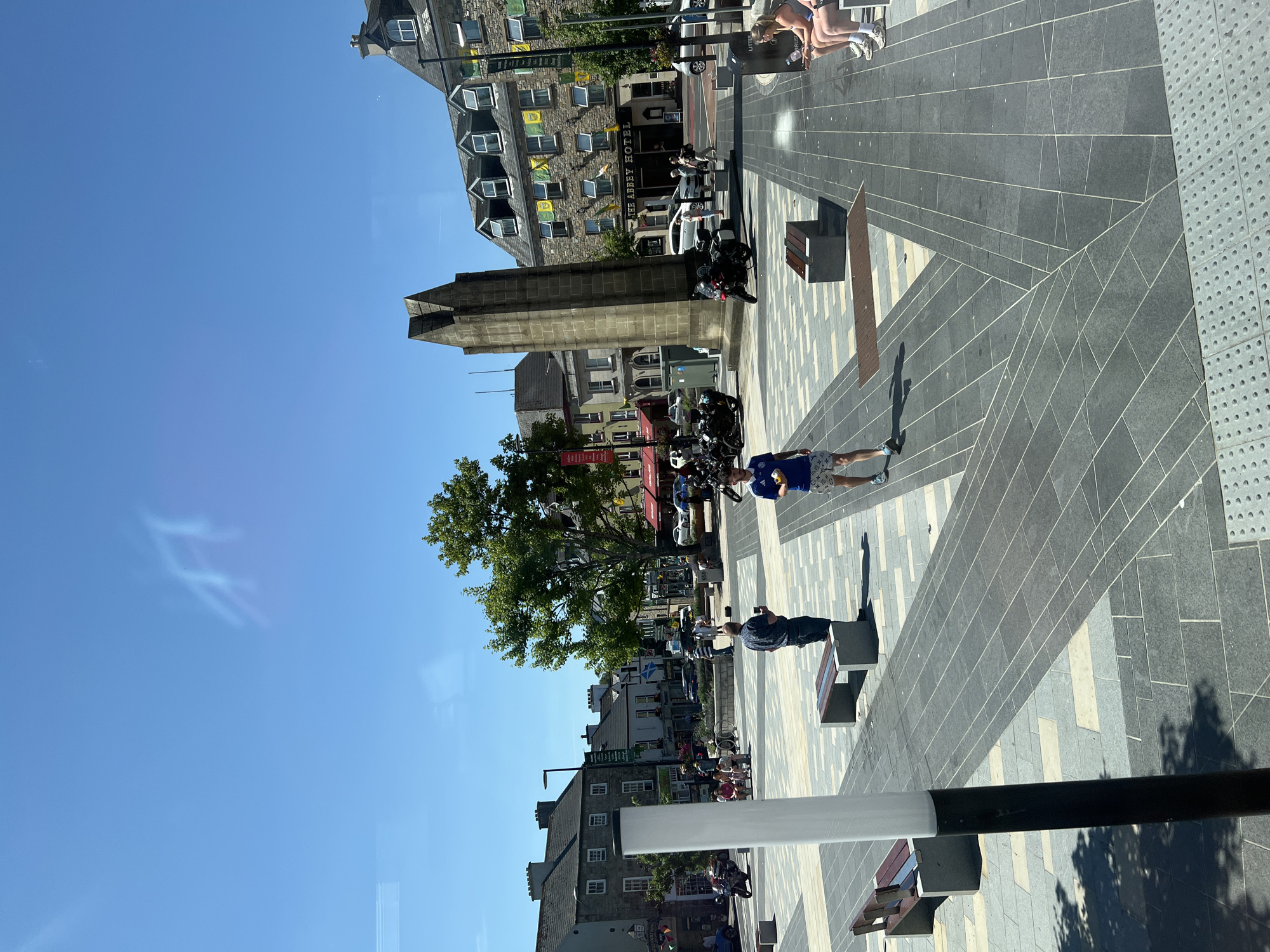 This is an outdoor, daytime photograph of a town square in Donegal, Ireland. In the foreground, a dark pole with a white cylindrical top is visible on the left. The square is paved with grey and lighter-colored rectangular tiles laid in a patterned design. Benches are placed throughout the square.
A tall, pointed stone monument stands prominently in the midground. To the right of the monument is a building with the sign "THE ABBEY HOTEL." The building has a stone facade and multiple windows with dormers on the roof. Flags, some yellow and green, are hanging from the windows.
Several people are present in the square. A man in a blue patterned shirt and dark shorts stands in the foreground, looking at his phone. Further back, a man in a blue and white checkered shirt and dark pants is holding a phone. A man in a blue t-shirt and patterned shorts is walking away from the camera. In the background, there are more people, some sitting on benches and others walking. Motorcycles are parked in various locations within the square.
Trees with green foliage are visible, and beyond the square, there are more buildings characteristic of a town center, with shops and signage. The sky is clear and blue. Reflections of the sky and possibly trees are visible on a surface in the upper left portion of the image.