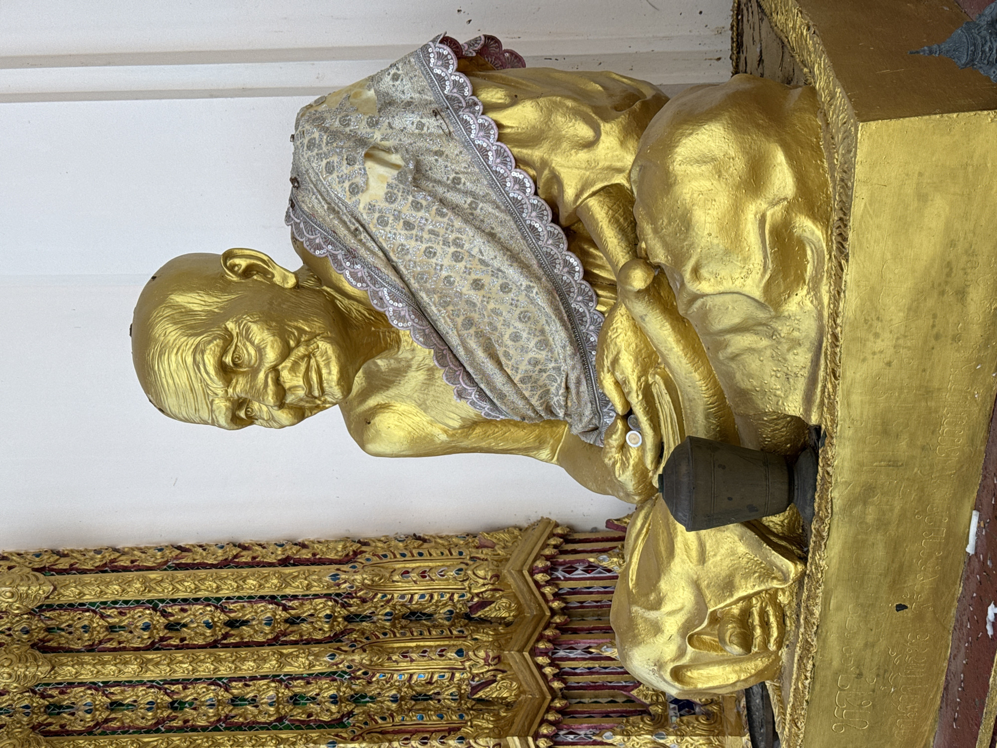A golden statue of a seated monk is the central subject of this image, positioned within what appears to be a temple or shrine in Ko Samui District, Thailand. The monk is depicted with his legs crossed and his hands resting on his lap, holding what appears to be a small, dark vessel. He is adorned with a light-colored, patterned cloth draped over his shoulder and chest, trimmed with a decorative border of small sequins and pink stitching. The statue's surface is highly reflective, showing some imperfections and subtle variations in its golden sheen.
To the left of the statue, a richly decorated golden architectural element is visible, featuring intricate carvings and inlaid colorful details, likely part of the temple's ornamentation. The background is a plain white wall. In the foreground, on the platform beneath the statue, a small, dark, urn-like object rests near the monk's hands. The base of the statue itself is also golden and appears to have inscriptions in an unfamiliar script. Scattered on the ground in front of the base are small white objects, possibly offerings or debris. The lighting suggests an outdoor or well-lit indoor environment. The overall scene evokes a sense of reverence and cultural significance associated with Buddhist art and architecture.