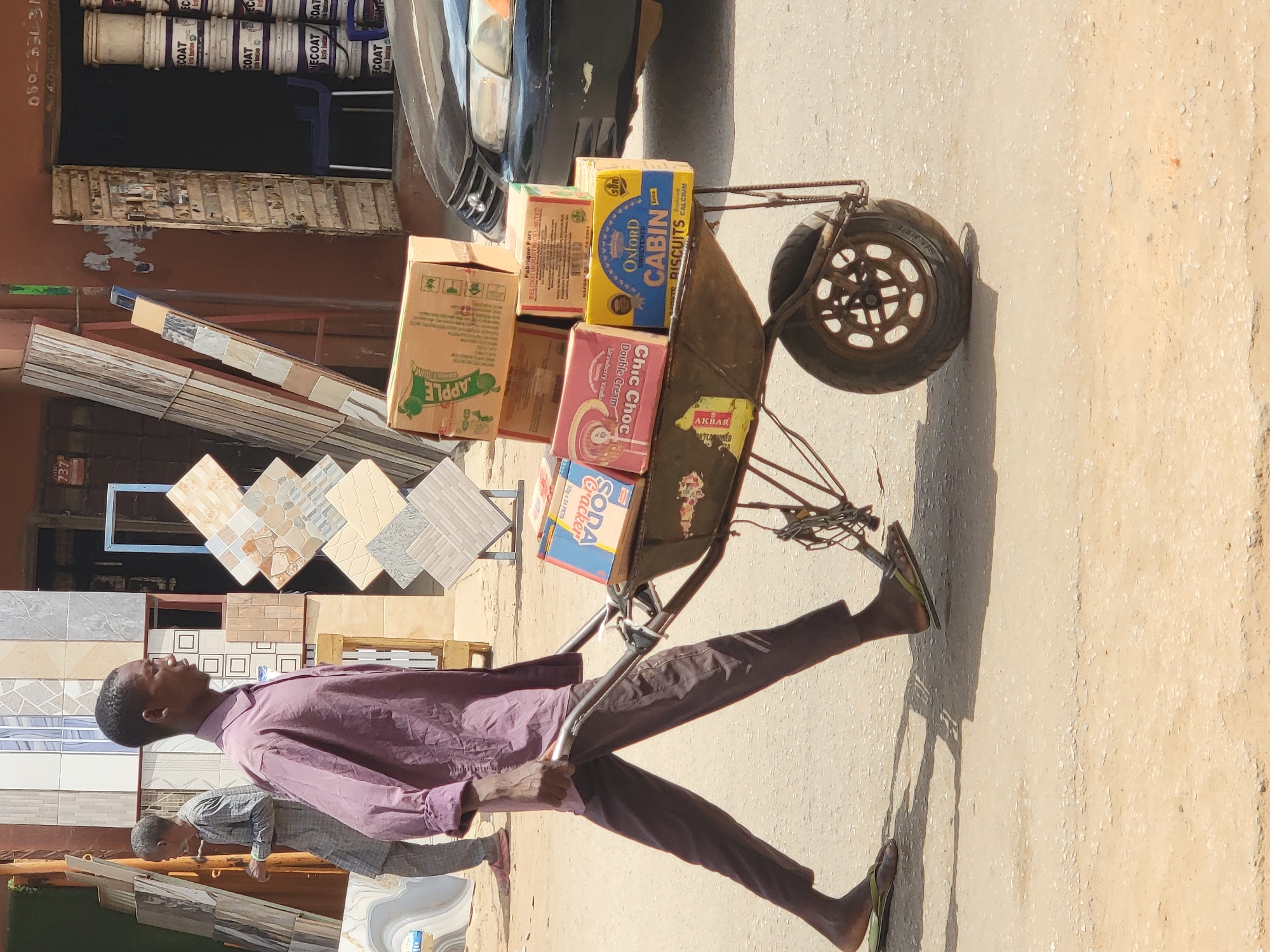 A man is pulling a rusty metal hand-drawn cart with a load of goods through a commercial district in Maiburiji, Nigeria. The cart is laden with boxes of Oxford Cabin biscuits, Chic Choc chocolate bars, Soda Crackers, and an “Apple” branded box of goods. The man is wearing a mauve shirt and brown pants, and sandals, and he is walking barefoot. He appears to be in transit and is heading towards a roadside vendor that sells tiles and some decorative products with paint buckets located in the background. Another person is also visible, carrying a load and assisting the man. A car is partially visible in the background.