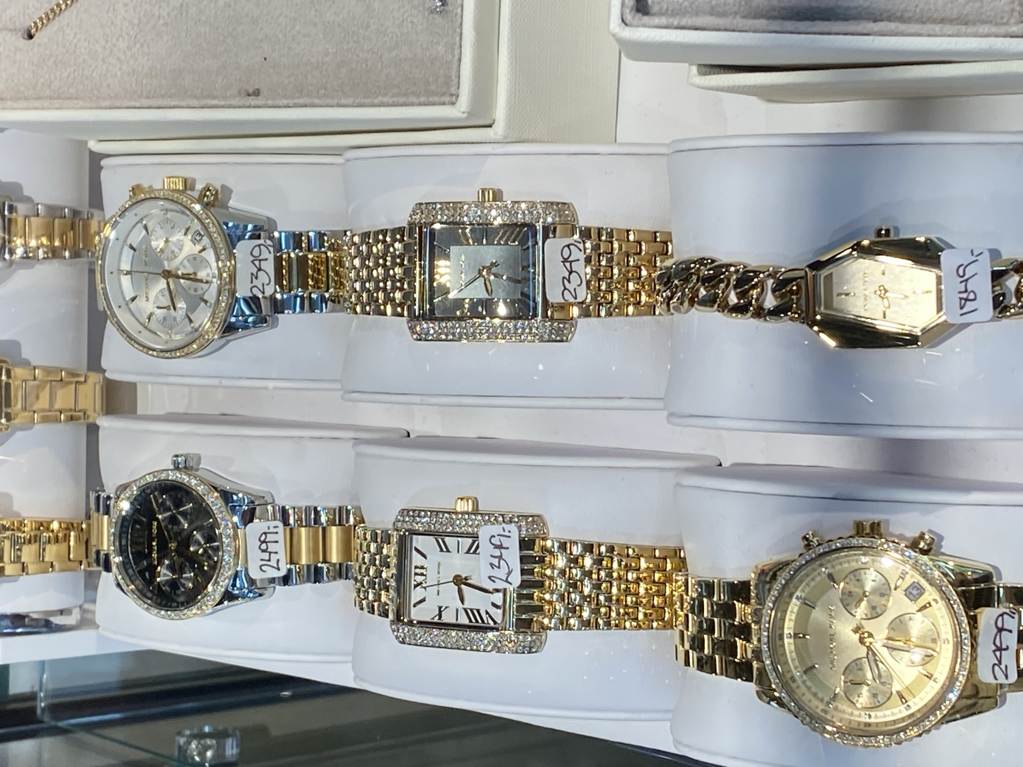 The image displays a close-up view of several gold-colored watches displayed on white stands. The watches vary in design, with some featuring diamond-encrusted bezels and others having more elaborate faces with multiple dials. Price tags are visible on some of the watches, indicating prices such as "2499,-", "2349,-", and "1849,-". The background is blurred, suggesting a retail environment such as a jewelry store. The watches are the primary focus, presented in a well-lit manner to highlight their details and craftsmanship. The overall impression is one of a high-end product display.