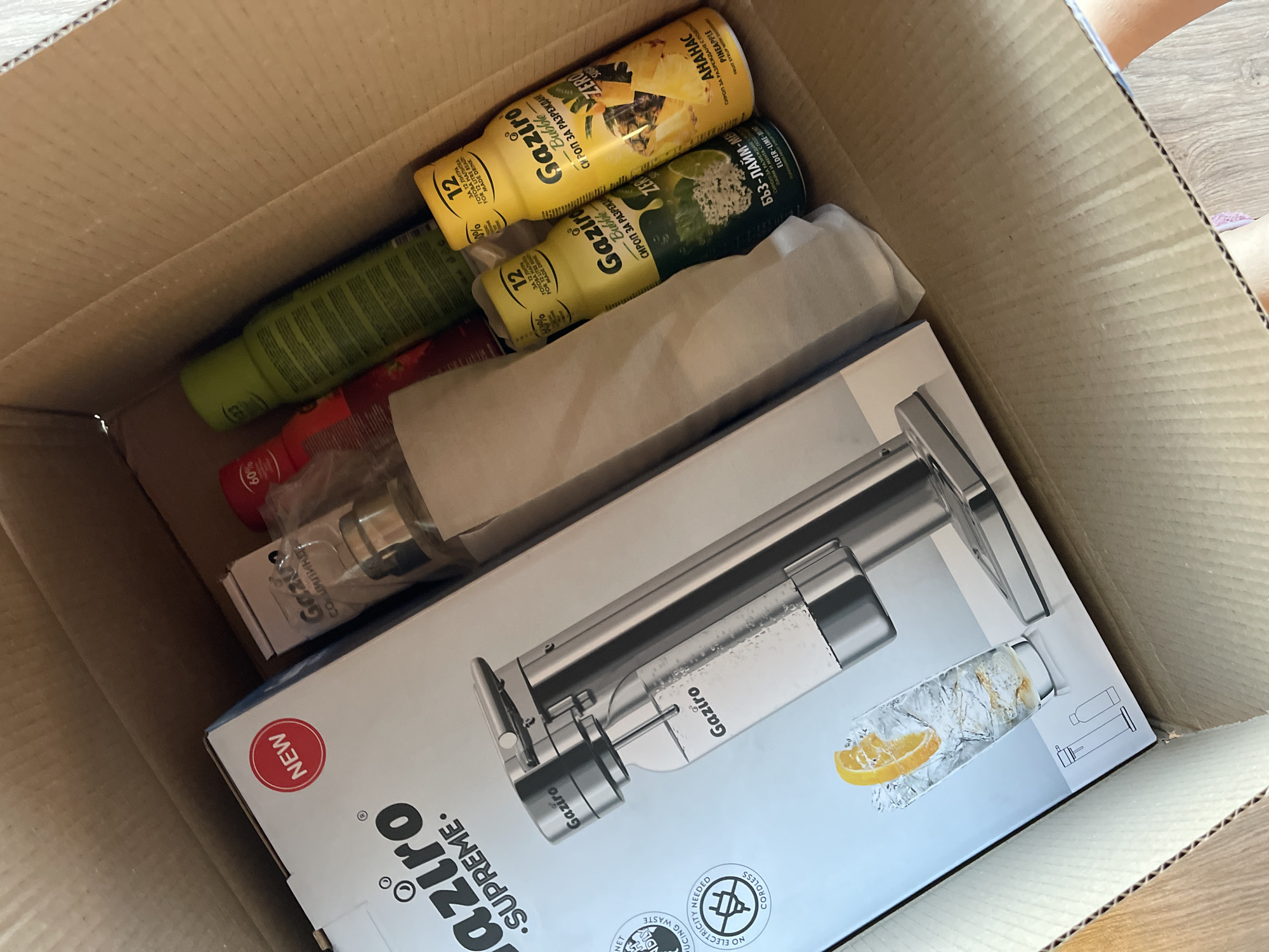 The image shows a cardboard box filled with several items, suggesting a recent delivery. Prominently featured is a product labeled "Gaziro Supreme", packaged in a white box with a depiction of a carbonated drink dispenser. Accompanying the dispenser are several bottles of Gaziro syrups in flavors like pineapple and elder-lime-mint, along with other bottles of unspecified contents. The scene appears to be indoors, with a portion of a wooden floor visible. The presence of Bulgarian text on the syrup bottles, such as "СИРОП ЗА РАЗРЕЖДАНЕ" and "ЗА 12 ЛИТРА", indicates that the box's contents are likely located in Bulgaria, with Sofia as a possible location given the product's availability in urban areas.
