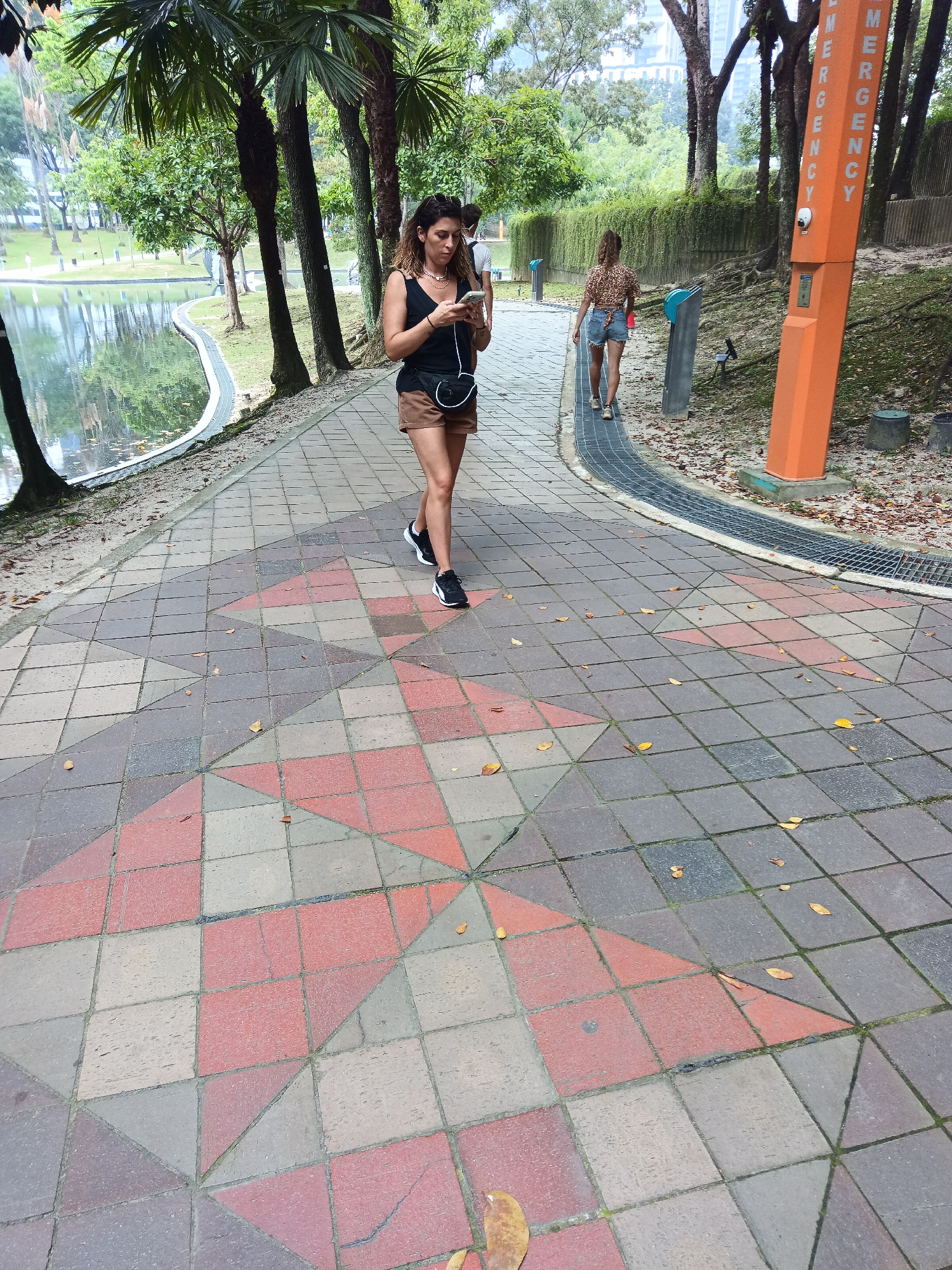 The image shows a woman walking on a paved pathway in a park with a body of water and trees. She is wearing a black tank top, brown shorts, and black sneakers. She is looking at her phone, which she is holding in her right hand. She has a black fanny pack across her chest. Behind her, another woman is walking away from the camera, wearing a patterned top and denim shorts. An orange emergency call box with the word "EMERGENCY" written vertically on it is visible to the right. The pathway is made of square tiles in various shades of red, pink, and grey, with a geometric pattern. The park appears to be in Kuala Lumpur, Malaysia, as suggested by the architecture and vegetation.