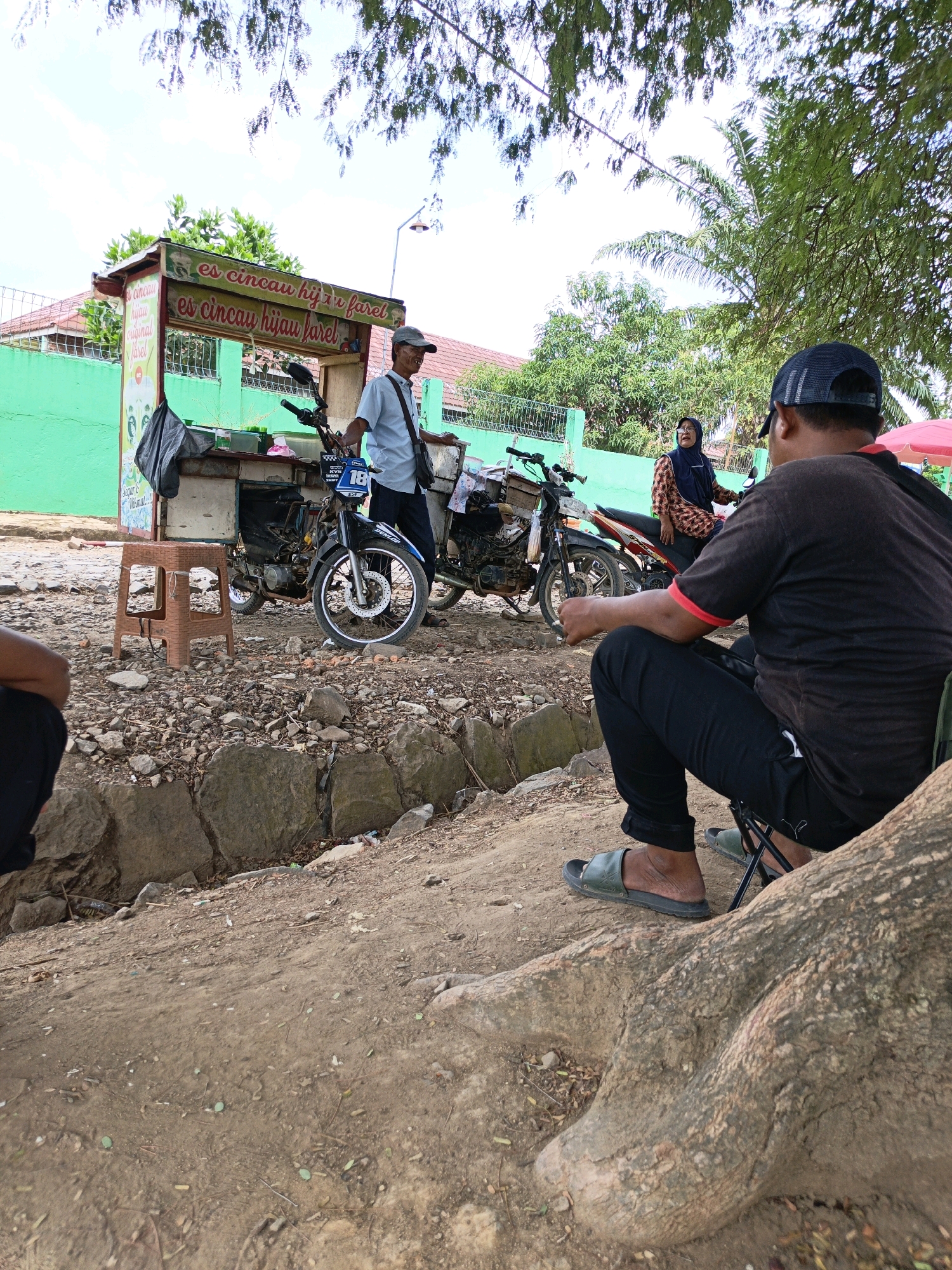 The image depicts an outdoor scene in the city of Gedung Nyapah, Indonesia. In the mid-ground, a food cart with the text "es cincau hijau farel" is set up, suggesting it sells "es cincau" (grass jelly drink). A man in a light blue shirt and a cap is standing by the cart, interacting with it. Two motorcycles are parked near the cart; one is a black and blue motorcycle with the number "18" on its front, and the other is a red and black motorcycle. A woman in a headscarf and patterned top is standing near the red and black motorcycle. In the foreground, a person is seated on a folding chair, facing away from the camera and towards the food cart and motorcycles. The ground is a mix of dirt, rocks, and tree roots, indicating a somewhat unpaved or natural environment. Trees and foliage are visible in the background, with a green wall and buildings also present, suggesting a residential or semi-urban setting. The lighting suggests it is daytime.