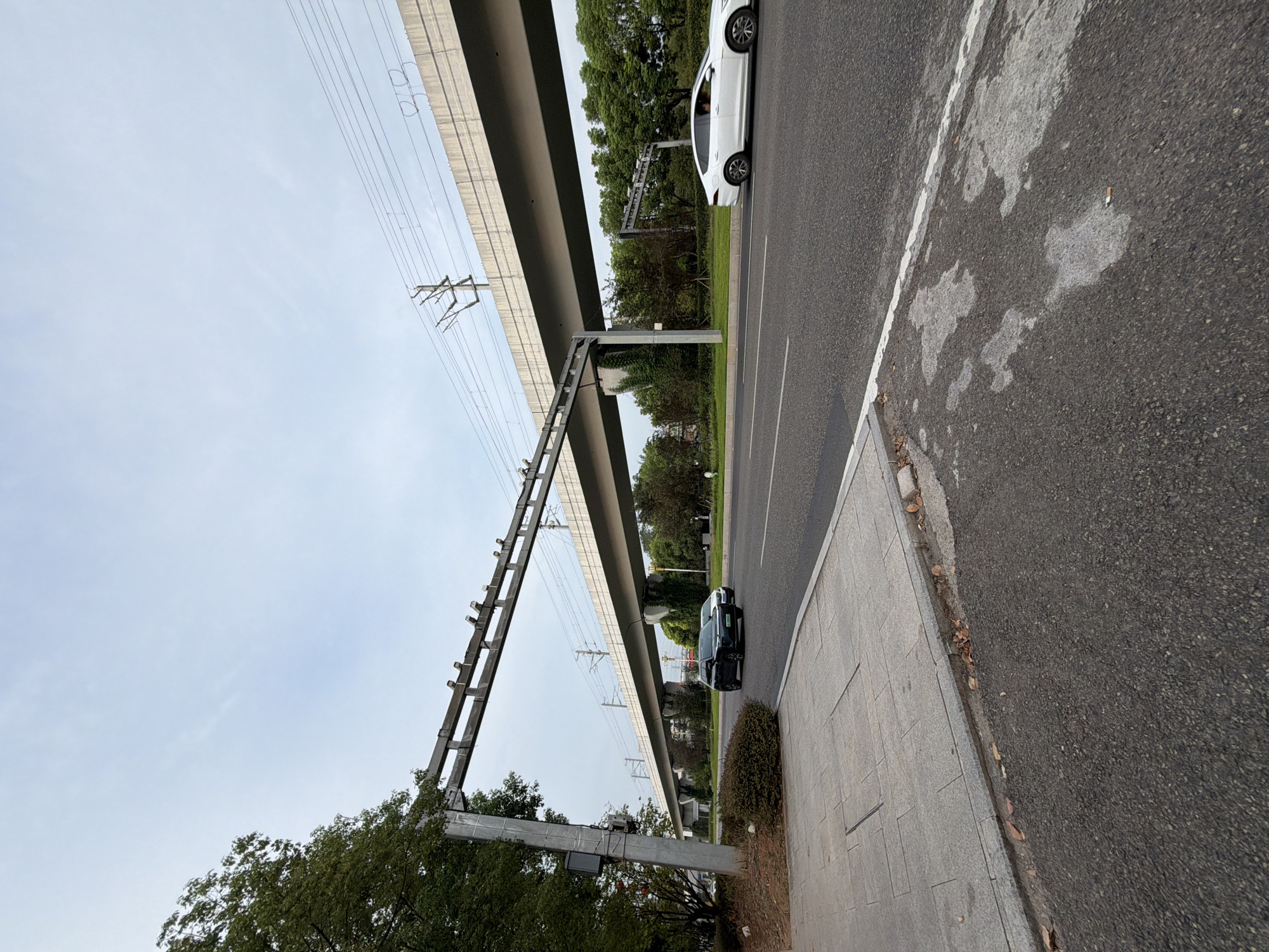 This image depicts a street scene in Jiangjin, China, featuring a modern elevated railway track supported by concrete pillars. The railway structure runs horizontally across the frame, with overhead power lines visible against a pale, overcast sky. Below the elevated track, a two-lane road stretches into the distance. On the right side of the road, a white sedan is in motion. On the left side, a dark-colored SUV is also moving, followed by another vehicle partially obscured. The roadside on the left includes a paved sidewalk and a strip of green vegetation, including a lush tree. The road surface appears to be asphalt, with some lighter patches and markings. The overall impression is one of urban infrastructure and daily transportation.
