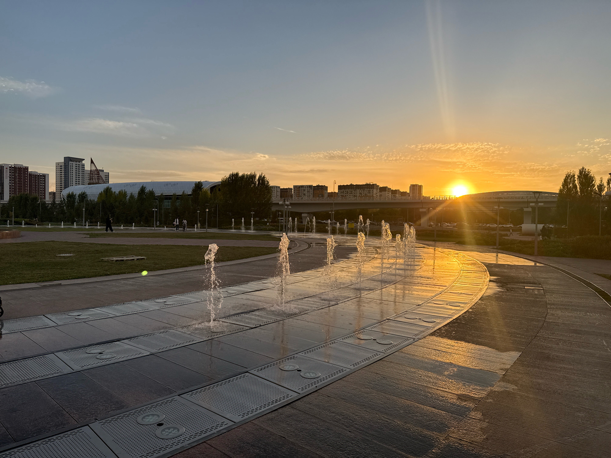 This image depicts a sunset over a modern urban park in Astana, Kazakhstan. The foreground features a decorative fountain with multiple water jets spraying upwards from a flat, tiled surface. The water reflects the warm, golden light of the setting sun, creating a shimmering effect on the wet pavement. In the middle ground, a grassy area is visible with a few scattered people walking and cycling. Beyond the park, a city skyline is apparent, dominated by tall residential buildings and a large, arched stadium with a white, undulating roof. A highway overpass with cars can be seen in the distance. The sky is clear with a few wispy clouds, and the sun is low on the horizon, casting long shadows and bathing the scene in a warm, orange glow. The overall impression is one of a vibrant and contemporary urban space at the end of a day.