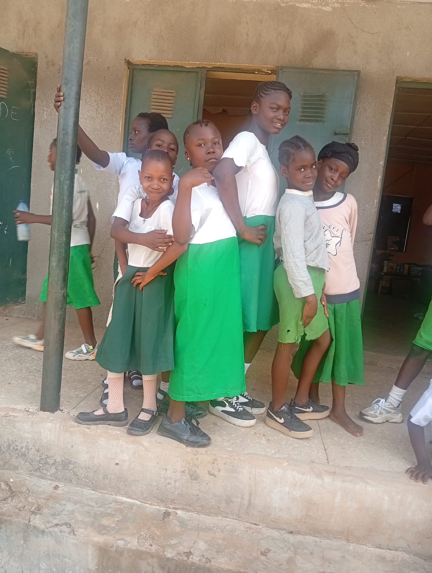 A group of seven schoolgirls, most of whom are wearing white shirts and green skirts, pose together on a concrete surface in front of a building. One girl wears a grey shirt and a torn green skirt, and another wears a pink shirt with a black head covering. Some of the girls are wearing shoes, while others are barefoot. A dark green chalkboard is partially visible on the left side of the frame, and there are green metal doors in the background. The scene is located in Narayi, Nigeria.