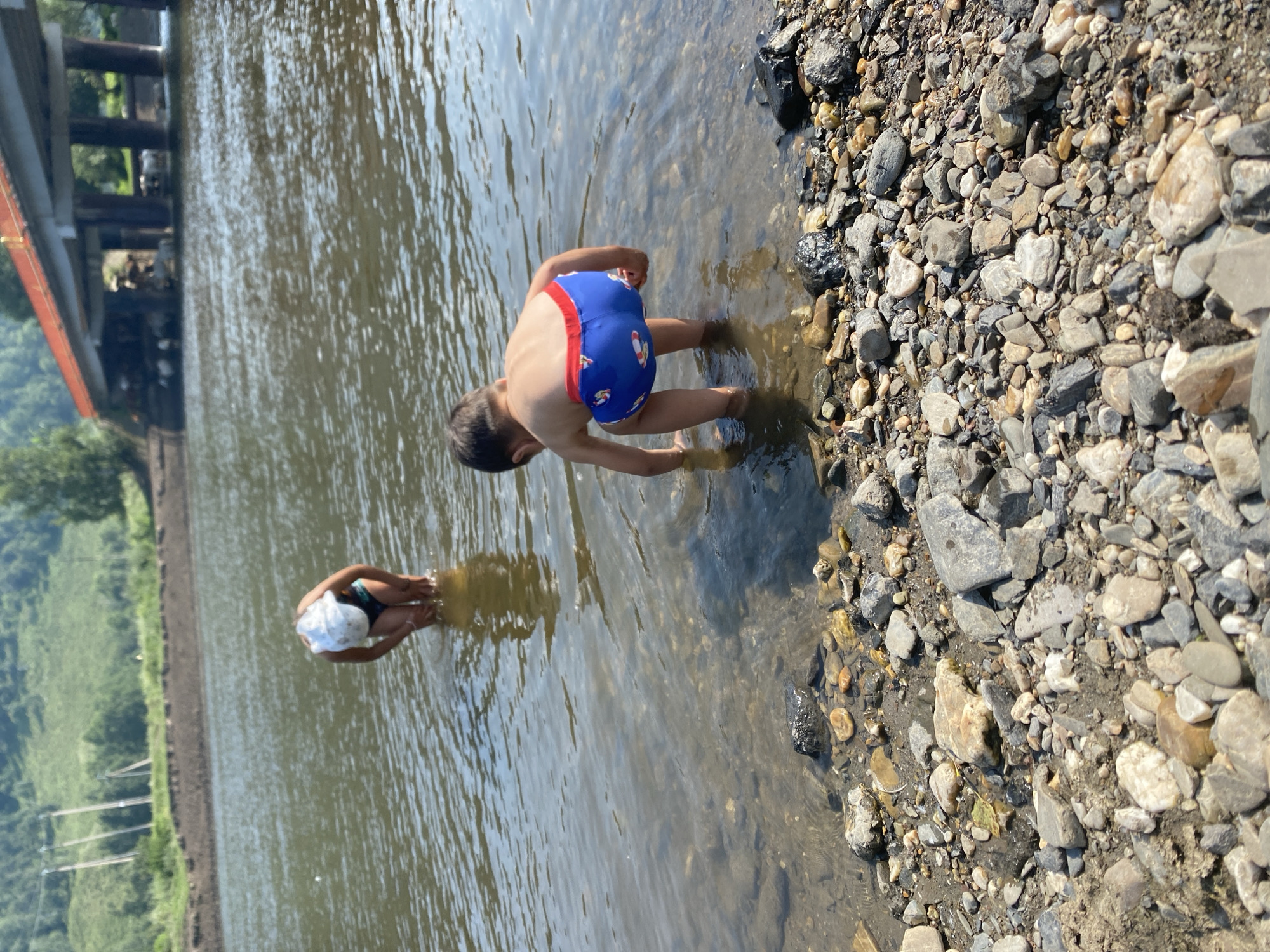 Two children are wading in the shallow waters of the Irtysh River, near the village of Maloarslangulovo, Russia. The boy in the foreground, facing away from the camera, wears blue swim trunks with a life preserver pattern. He is bent over, reaching into the water with his hands. Behind him, a child wearing a white hat and dark swimsuit is also in the water, facing away from the camera and bending over. The riverbank in the foreground is covered in pebbles and rocks. In the background, a bridge spans the river, supported by concrete pillars. Lush green hills with scattered trees rise beyond the river. The water appears murky, and the sky is bright and sunny.