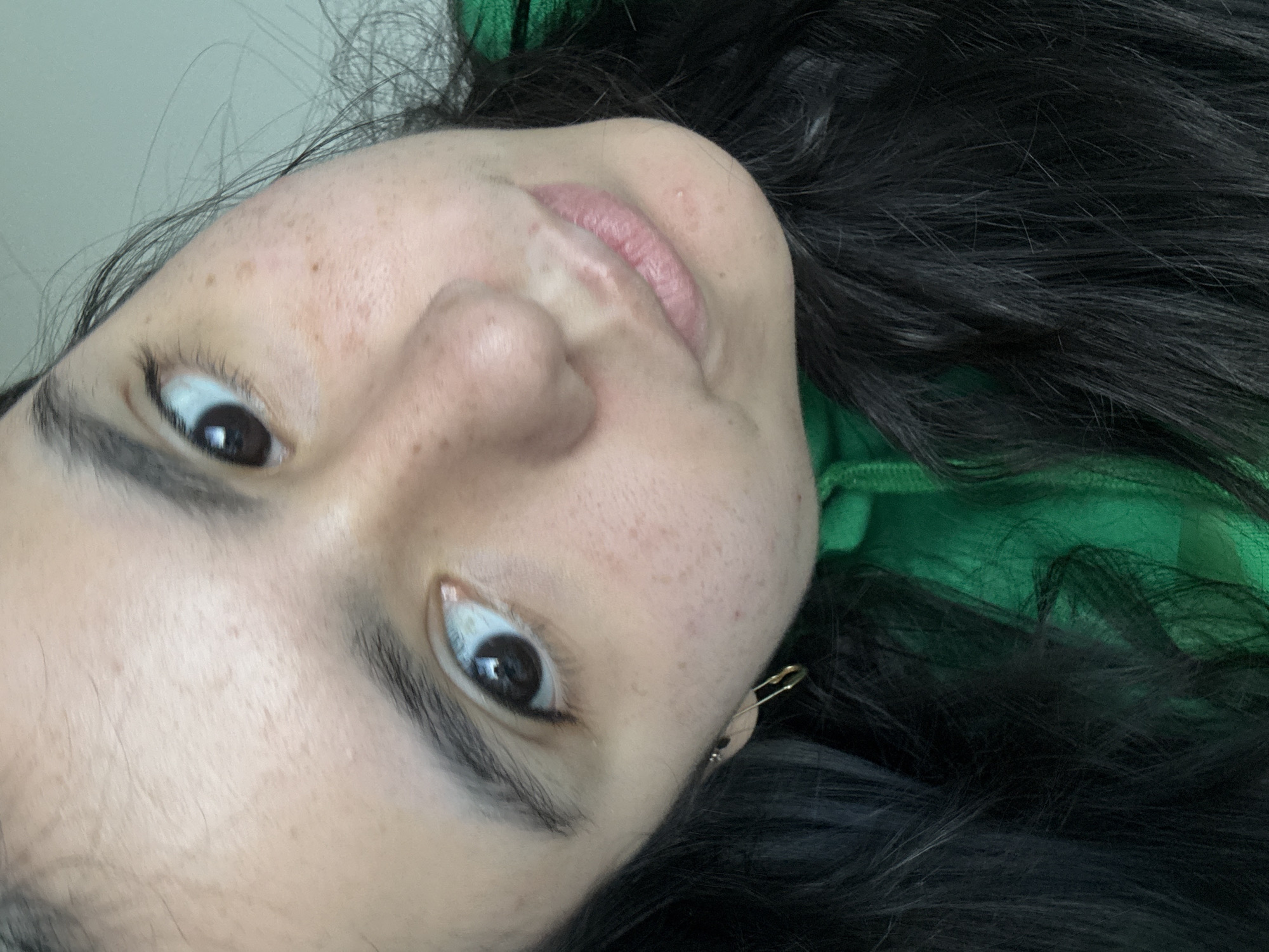 A close-up, angled selfie shows a person with dark hair and fair skin. The subject is wearing a green top and a gold safety pin is visible near their left ear. The background is a plain white wall. There is no contextual information in the image to determine a location in Austin, United States of America, or any other specific location.