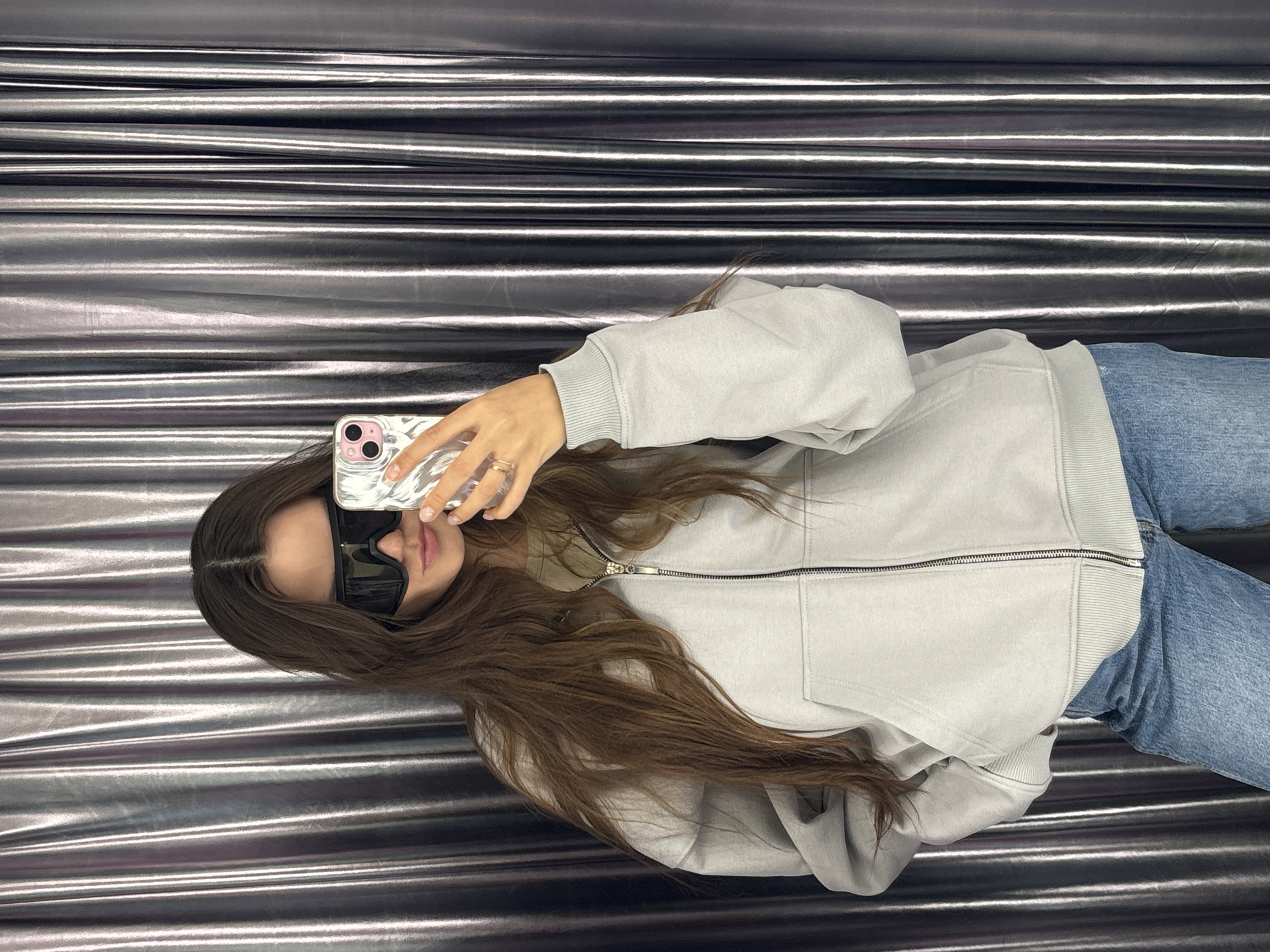 A young woman is lying on a metallic surface, likely a bed, taking a selfie. She is wearing a light grey zip-up hoodie, blue jeans, and large black sunglasses. Her long brown hair is spread out around her head. She is holding a smartphone with a clear case and a distinctive dual-lens camera setup, possibly an iPhone. The background is a glossy, reflective material with horizontal striations. There is no information to locate this scene in the city of Krasnoyarsk, Russia.