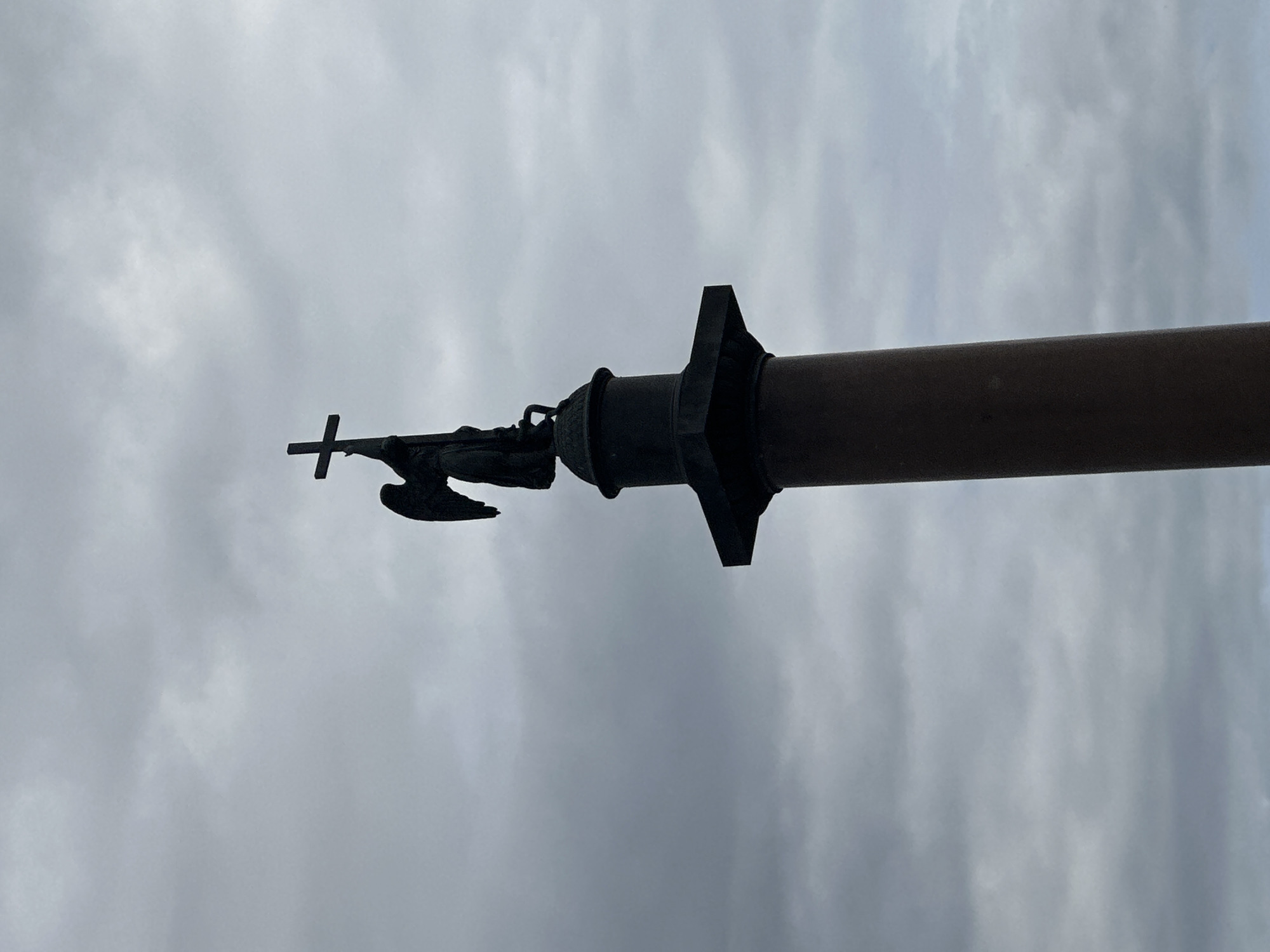 The image is a low-angle, close-up shot of a dark, cylindrical object, likely a monument or structure, against a cloudy, overcast sky. A dark, stylized winged figure is visible on the left side of the frame, positioned atop the cylindrical object and holding a cross. The figure and cross are silhouetted against the sky. The cylindrical object appears to be made of metal and has a textured surface, with a flared or decorative element near the figure. The sky is filled with diffused light, suggesting it is daytime, possibly late afternoon or a very cloudy day. There is no visible text in the image. The overall mood is somber and majestic, emphasizing the scale and imposing nature of the monument.