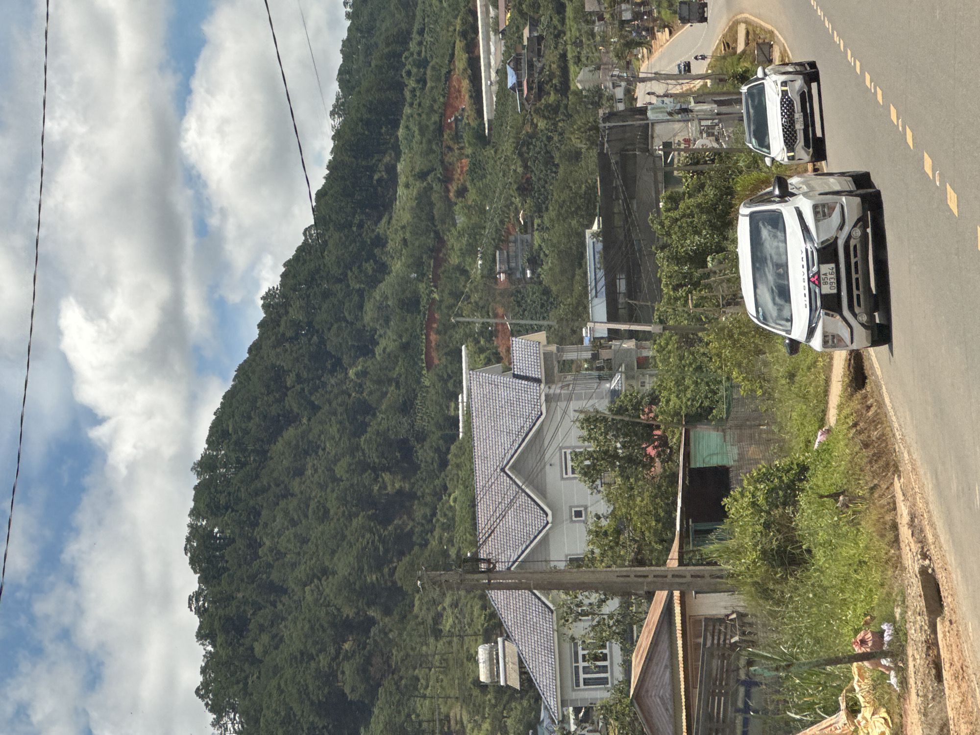 The image depicts a bright, daytime scene on a road in Thôn 1, Vietnam. A white Mitsubishi Xpander is in the foreground, driving on the right side of the paved road. A white Hyundai Santa Fe is behind it, also on the right side of the road. The road curves to the right, leading uphill. On the left side of the road, there is a grassy embankment with some vegetation and a large, light-colored house with a gray tiled roof. Further up the hill, more houses and buildings are visible, interspersed with dense green trees. The background is dominated by a heavily forested mountain slope under a partially cloudy sky. The overall impression is a rural or semi-rural residential area in a hilly region.
