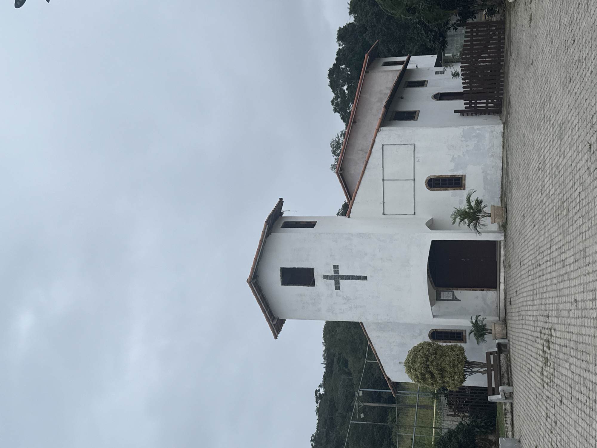 An exterior, eye-level, full shot captures a small white church with a bell tower against an overcast sky. The church has a red-tiled roof and a cross embedded in the facade of the bell tower, which has a dark rectangular opening at the top. The main body of the church features several arched windows and a large, dark double door. Potted plants and a stone bench with a wooden seat are situated in front of the church on a brick-paved courtyard. A wooden fence encloses a portion of the property to the right. The background is filled with lush green trees and foliage, suggesting a semi-rural or suburban setting. The image appears to be a photograph of a church located in the city of São Pedro Da Aldeia, Brazil.