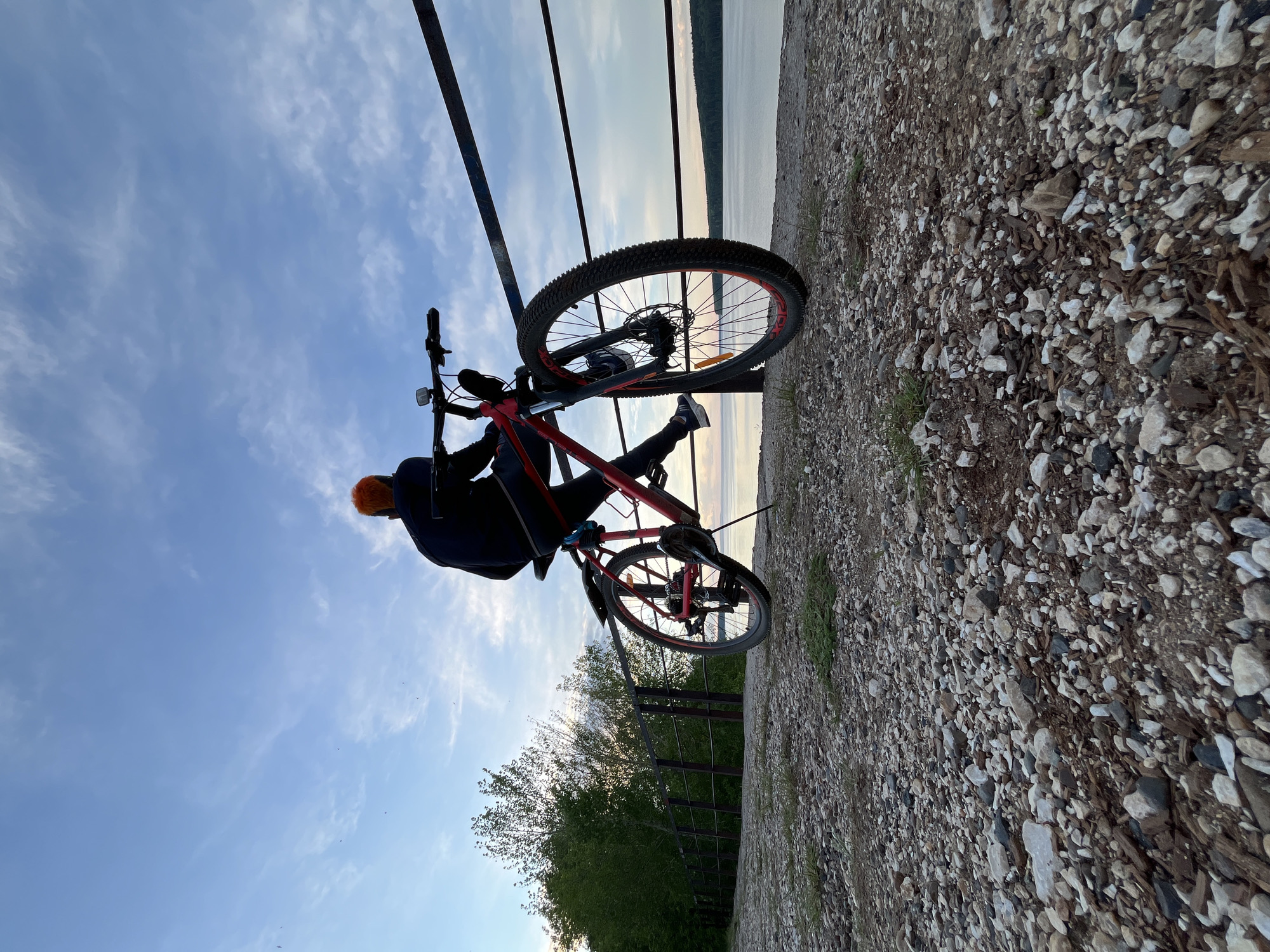 A man in a dark jacket and orange helmet sits on a red bicycle on a gravel path. The bicycle is leaning against a metal railing. Beyond the railing, there is a body of water and the horizon, which suggests the location may be the embankment in the city of Chaikovsky, Russia. There are green trees visible behind the railing on the left side of the frame. The sky is blue with scattered clouds. The gravel path is composed of light and dark colored stones and pebbles.