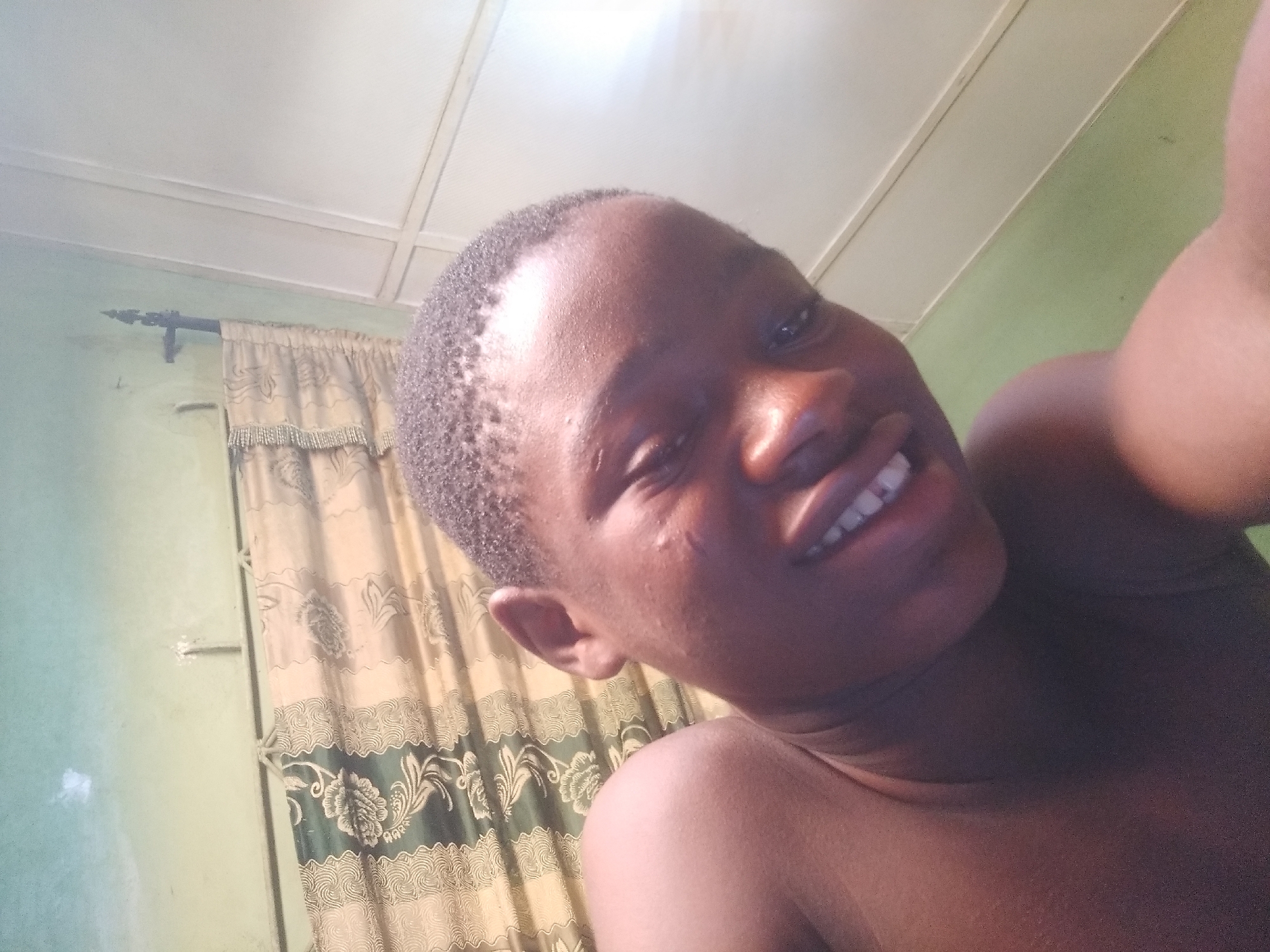 A close-up selfie shows a young person with dark skin and short, dark hair, smiling broadly at the camera. Their face is angled upwards, with bright light illuminating their features. The subject appears cheerful and relaxed.
The background reveals an indoor setting. A light green wall is visible to the left, adorned with a mounted curtain rod. Hanging from the rod is a patterned curtain featuring ornate floral or abstract designs in shades of beige, gold, and dark green or blue. Above, a white paneled ceiling completes the room's interior. The lighting is bright throughout the scene, suggesting either daylight or strong artificial illumination. No discernible text is visible in the image.