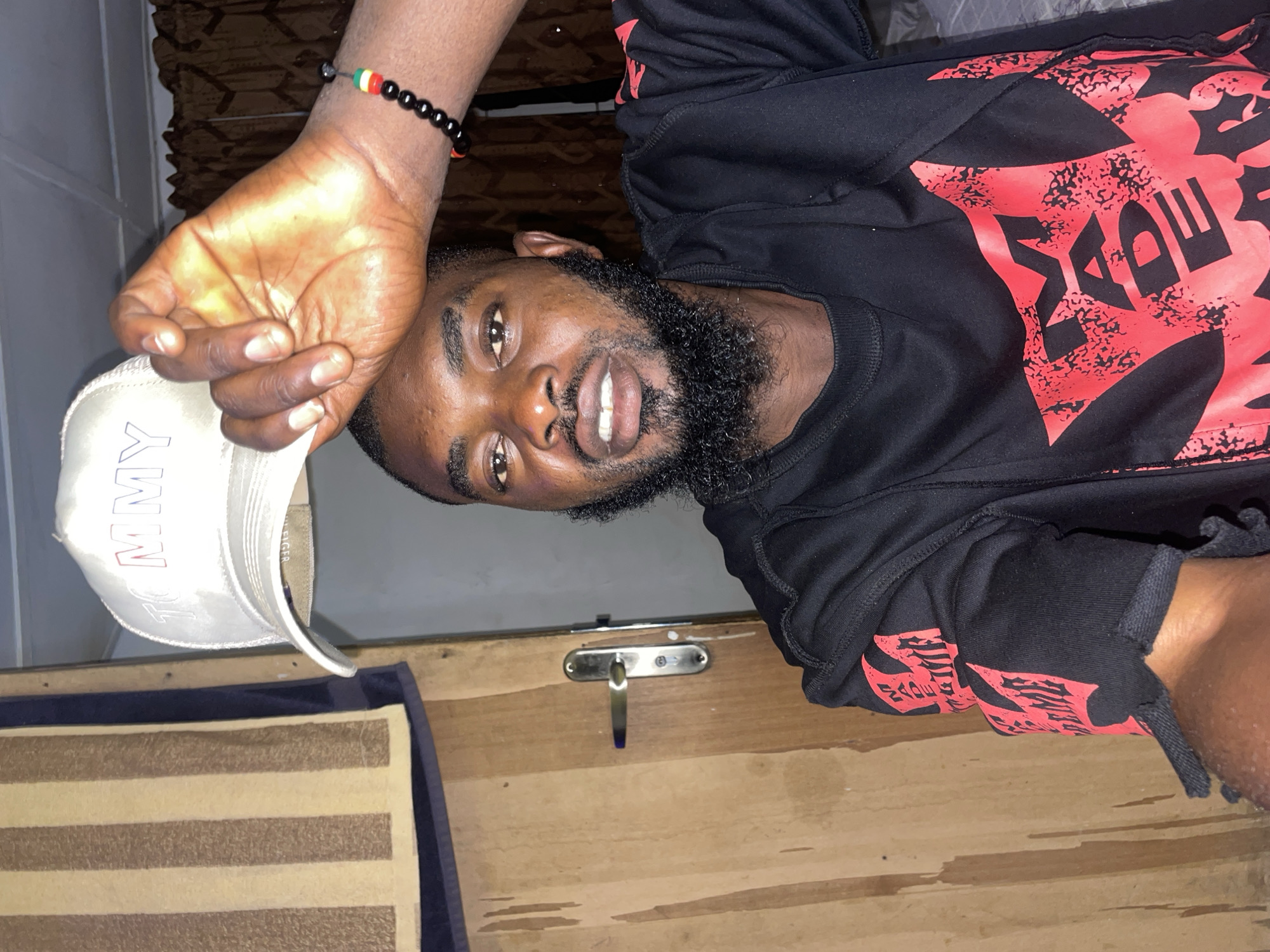 A young Black man with a beard is posing for a selfie indoors, holding a white baseball cap with "TOMMY" written on it over his head with his right hand. He is wearing a black t-shirt with red graphics. A beaded bracelet with red, yellow, and green beads is visible on his wrist. He is standing in front of a wooden door with a silver doorknob and a striped towel hanging to his left. The background consists of a patterned curtain. The scene appears to be within a residential setting in Jos, Nigeria.