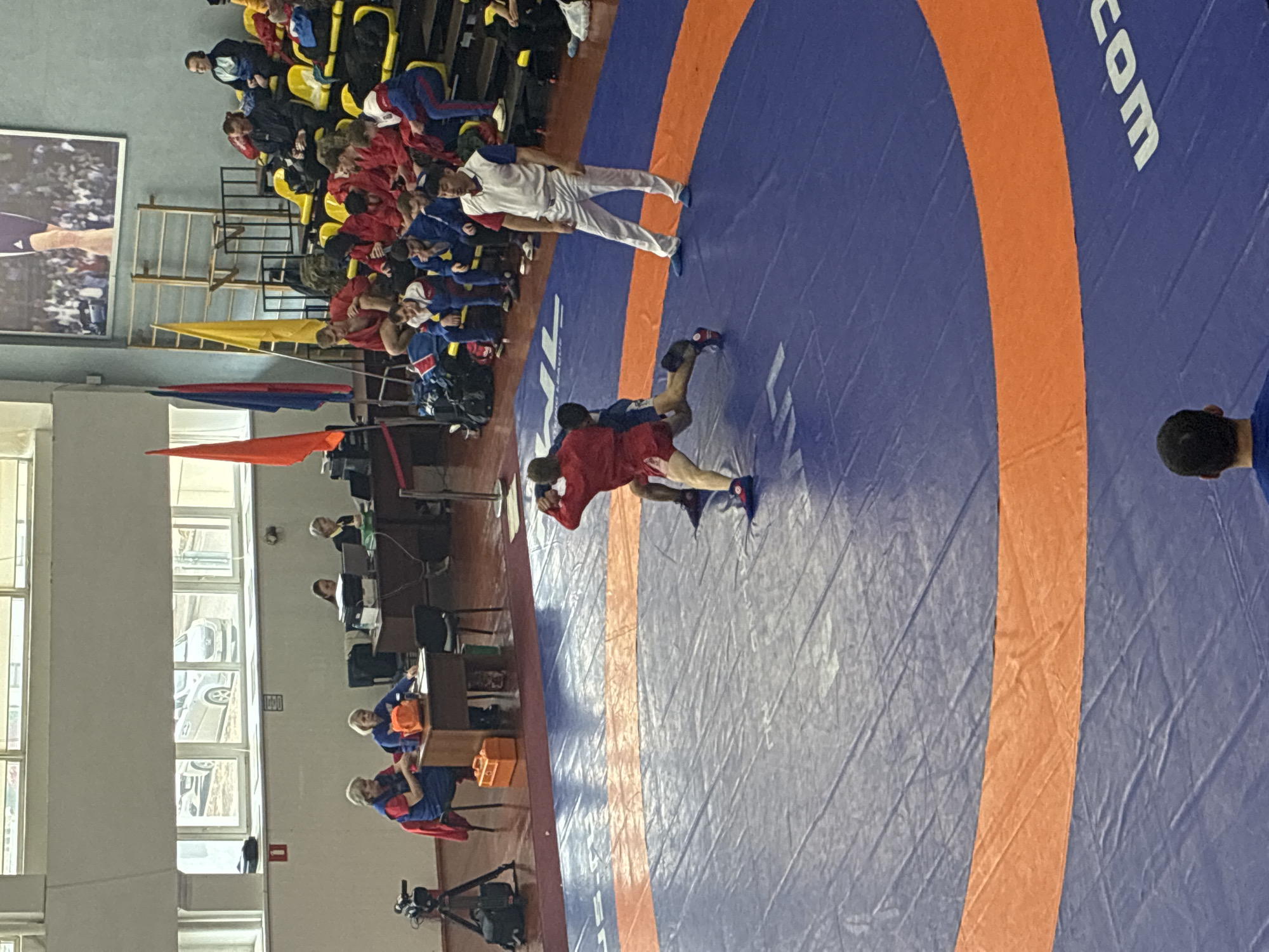The image depicts a wrestling match in progress on a blue mat with an orange border, possibly indoors, with spectators in the background. One wrestler wears a red uniform, while the other is in blue. An official in a white uniform oversees the match. To the side, several individuals are seated at tables, possibly scoring or judging the event. Flags and seating are also visible. The location appears to be a gymnasium or similar sports venue. The location is in the city of Unknown, Unknown.