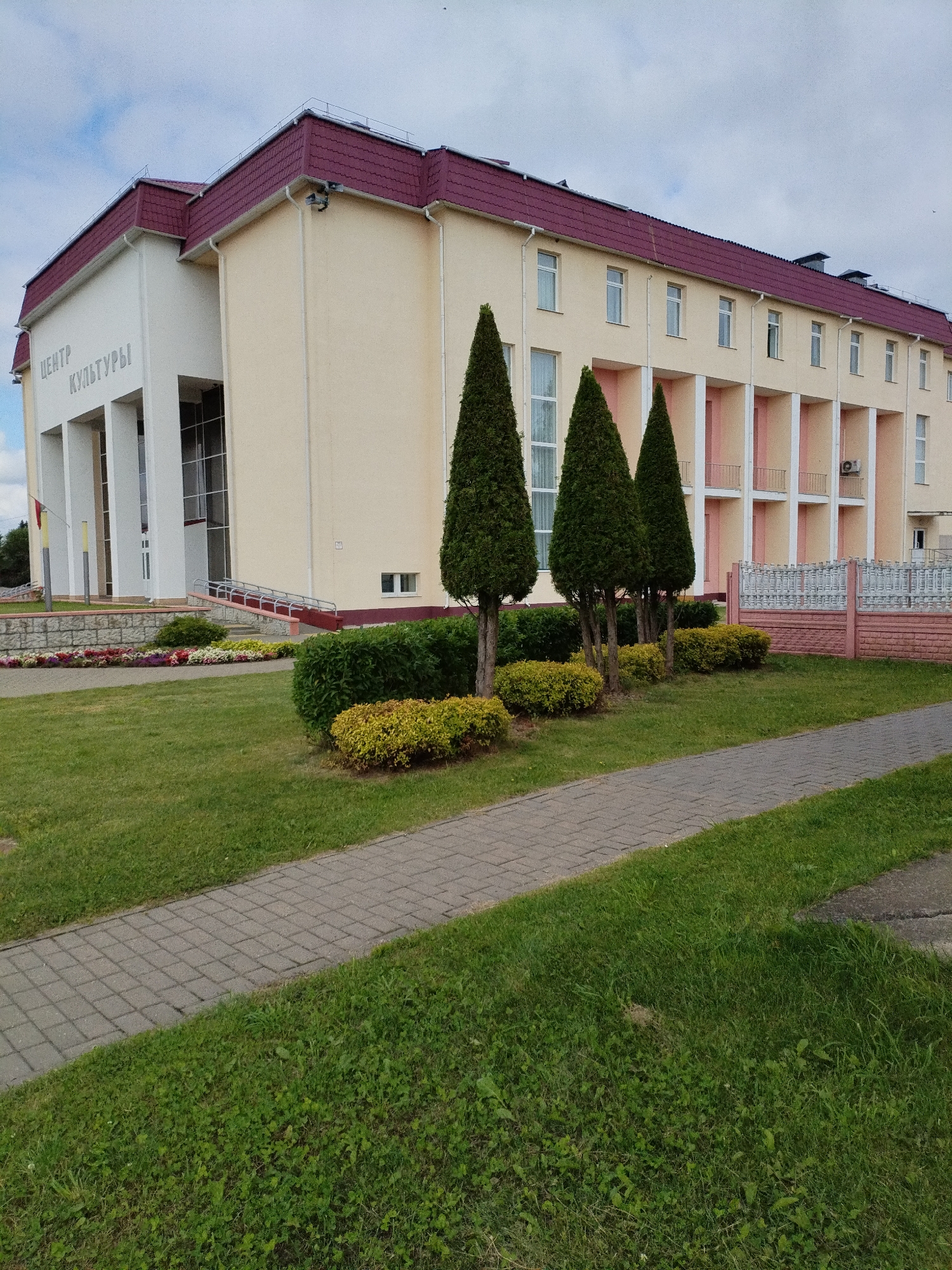 This is a full, eye-level, exterior, medium-shot photograph of the building identified by the signage as "ЦЕНТР КУЛЬТУРЫ" (Center of Culture) in Bialynichy, Belarus. The building is a light yellow, two-story structure with a prominent burgundy red roof. The left side of the building features a white facade with large windows and columns, and the words "ЦЕНТР КУЛЬТУРЫ" are emblazoned on this section in white lettering. The right side of the building has a repeated pattern of windows and pink balconies supported by cream-colored columns. There are three tall, conical evergreen trees standing in front of the building, with lower, manicured green and yellow bushes surrounding them. A paved walkway made of gray rectangular bricks runs diagonally across the lower right portion of the image, bordered by a well-maintained green lawn. In the background, a white ornamental fence with pink accents is visible. The sky above is partly cloudy with a few birds flying. The overall scene depicts a public building with a pleasant, landscaped exterior.