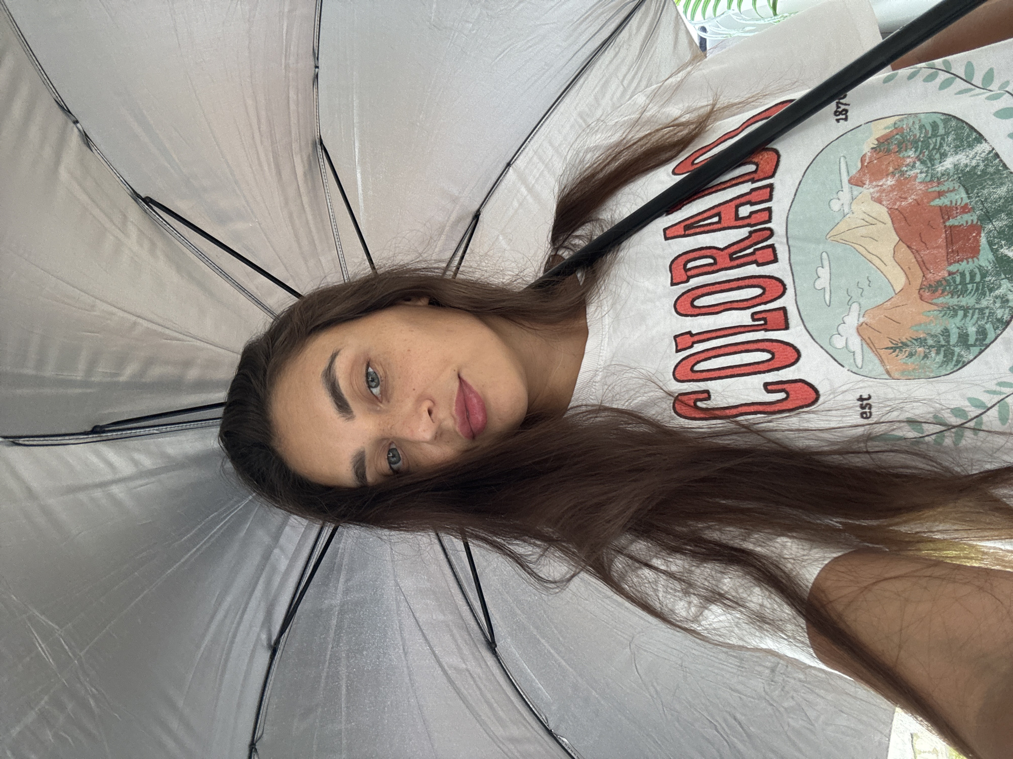A woman with light skin and long brown hair is lying down and looking directly at the camera. She is positioned underneath a large, open, silver umbrella. She is wearing a white t-shirt with a circular design featuring the word "COLORADO" in red and an image of mountains and trees. The background is indistinct, but there are hints of foliage. The location, Pa Khlok, Thailand, is not discernible from the visual information.