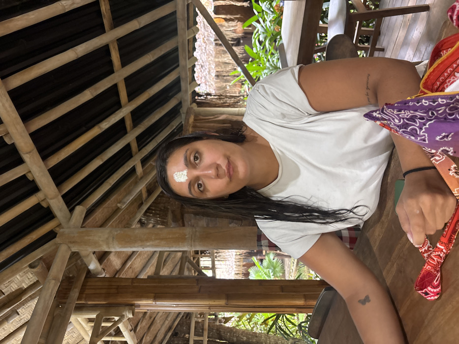 A single individual is seated at a wooden table within an open-air structure in Punggul, Indonesia. The person, appearing to be an adult female with dark, wet hair, faces the camera with a neutral expression. A white substance is present on her forehead. She wears a plain white short-sleeved t-shirt. A small dark tattoo, resembling a butterfly, is visible on her left forearm, and text, partially legible as "pour la personne," is tattooed on her right bicep/tricep area. Her hands rest on the table, near a folded collection of patterned cloths or bandanas in red, orange, and purple hues. A green object, possibly a smartphone, is partially visible on the table. The structure's framework consists primarily of bamboo poles, forming columns, beams, and roof supports. The roof appears to be covered with dark, possibly corrugated material. Lush green foliage and other wooden tables and chairs are visible in the background, suggesting a tropical outdoor or semi-outdoor setting.