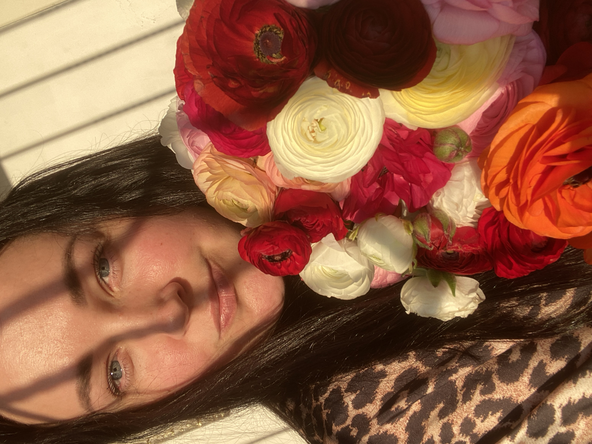 A woman is seen in a close-up, face-forward view, with a crown of ranunculus flowers in various colors atop her head. The flowers include shades of red, white, yellow, pink, and orange. The woman has fair skin, dark hair, and light-colored eyes. Shadows fall across her face, and she is wearing a patterned top with what appears to be a leopard print design. The background appears to be a white surface with shadows, possibly a wall or floor with light streaming through blinds. There is no clear indication of this photo being taken in Sochi, Russia.