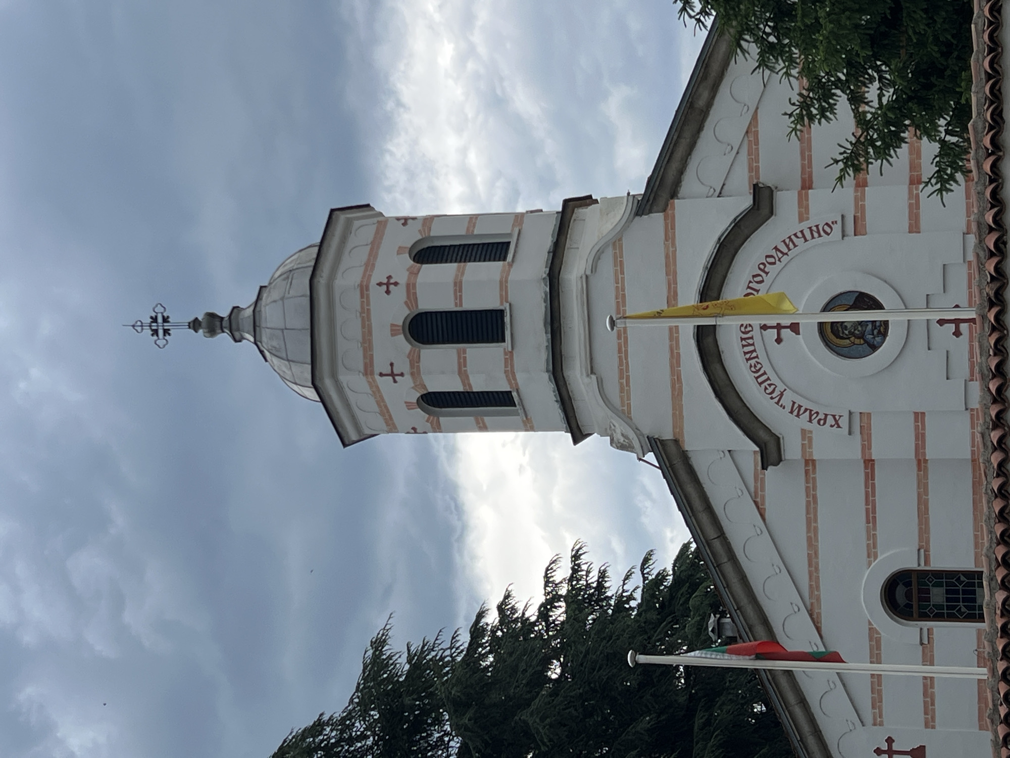 The image captures an upward view of the architectural details of a church, likely situated in Kazanlak, Bulgaria. The church is characterized by a white facade with vertical stripes of red brick design. A prominent feature is the tower, topped with a dome and cross. The tower has several arched windows.
Red Cyrillic lettering is visible on the building’s facade near the dome. Flags are flying from poles attached to the building. One flag is yellow. The other is the Bulgarian flag. Trees with green foliage frame the bottom of the image, providing a natural contrast to the building. The sky is overcast.