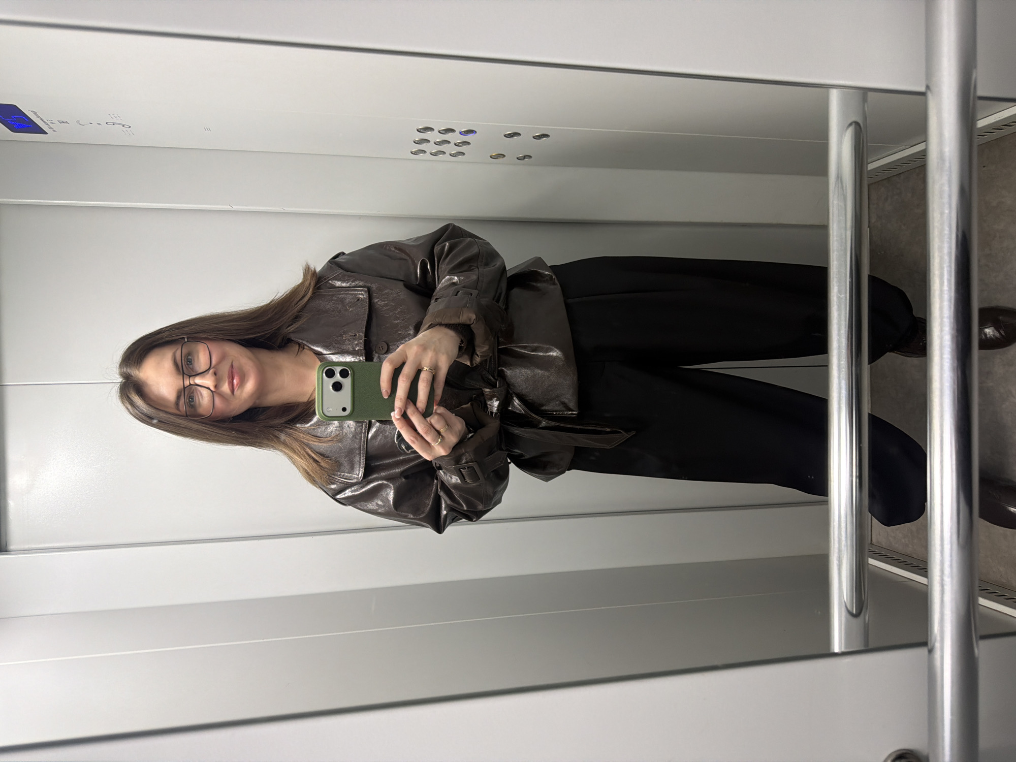 A woman is taking a selfie in what appears to be an elevator. She is wearing glasses, a brown leather jacket, and black pants. She is holding a green smartphone. The elevator has white walls, a control panel on the ceiling, and a handrail. The floor is visible in the reflection, showing a gray surface and brown shoes. It is not possible to confirm the location as Chaikovsky, Russia from the image.